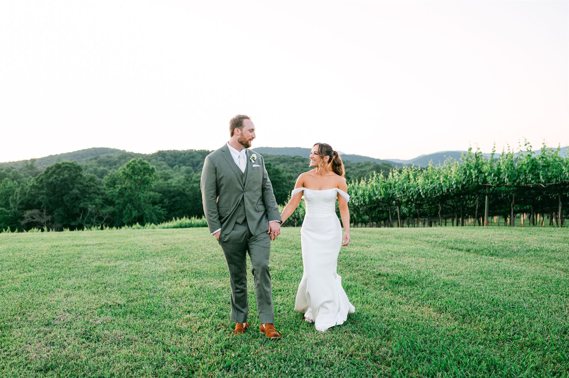 Bride and groom holding hands, smiling, walking in a field with vineyard in background.