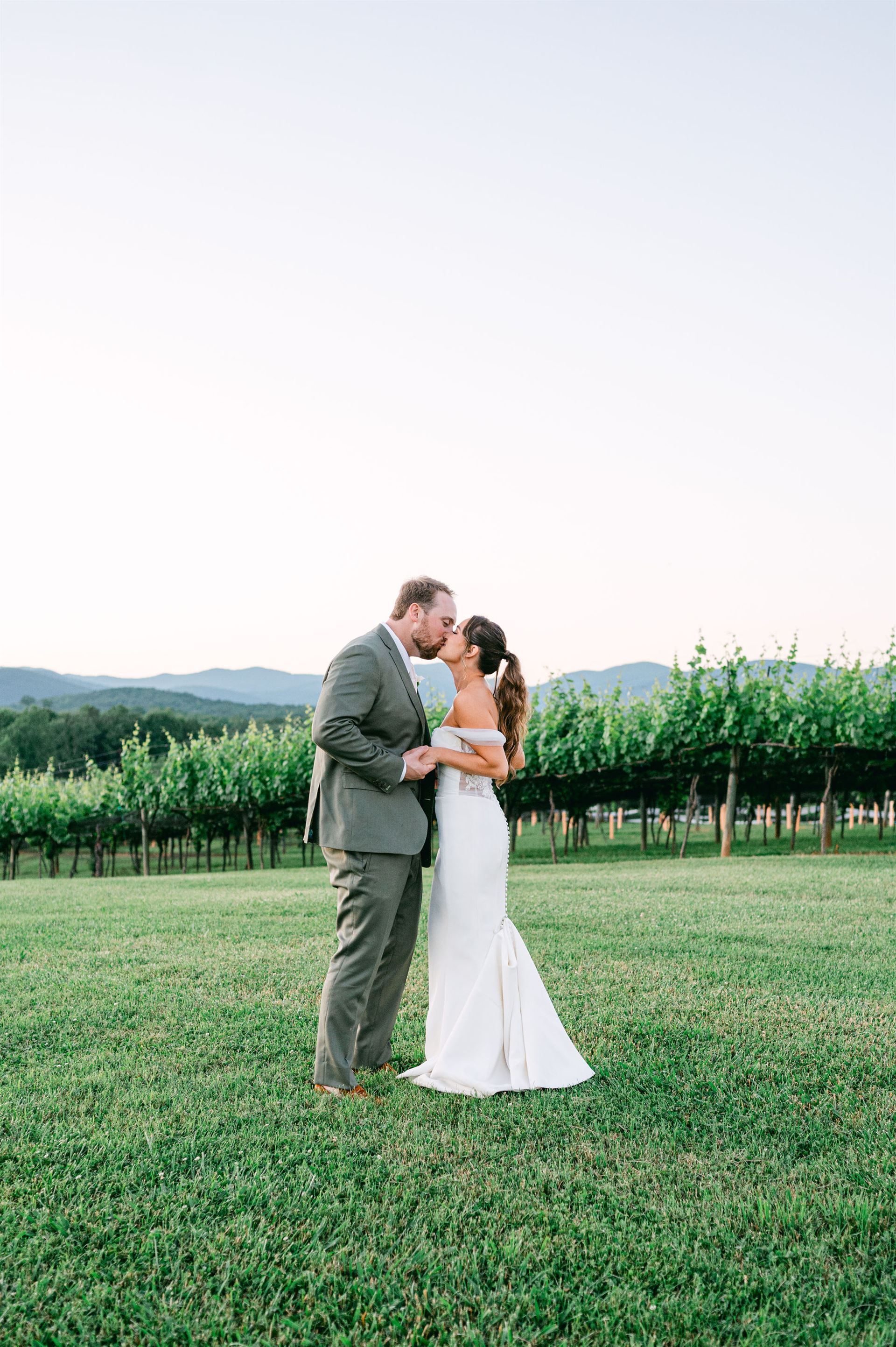 Couple kissing, standing in a vineyard. The groom wears a suit. The bride, a white gown. Green vines and mountains in the background.