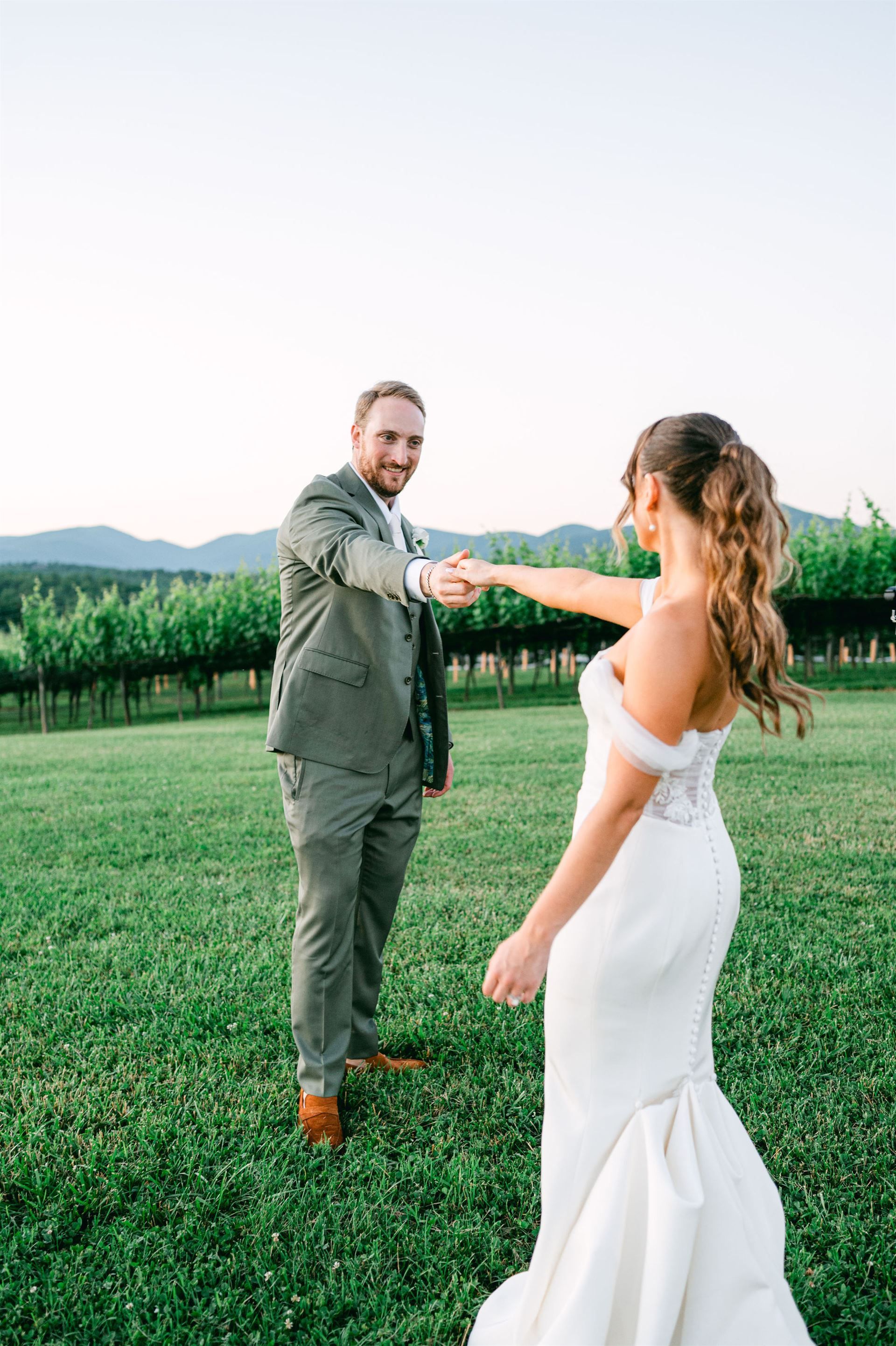 Bride and groom holding hands; groom gazes at camera, wearing green suit; bride in white gown; field, vineyard background.