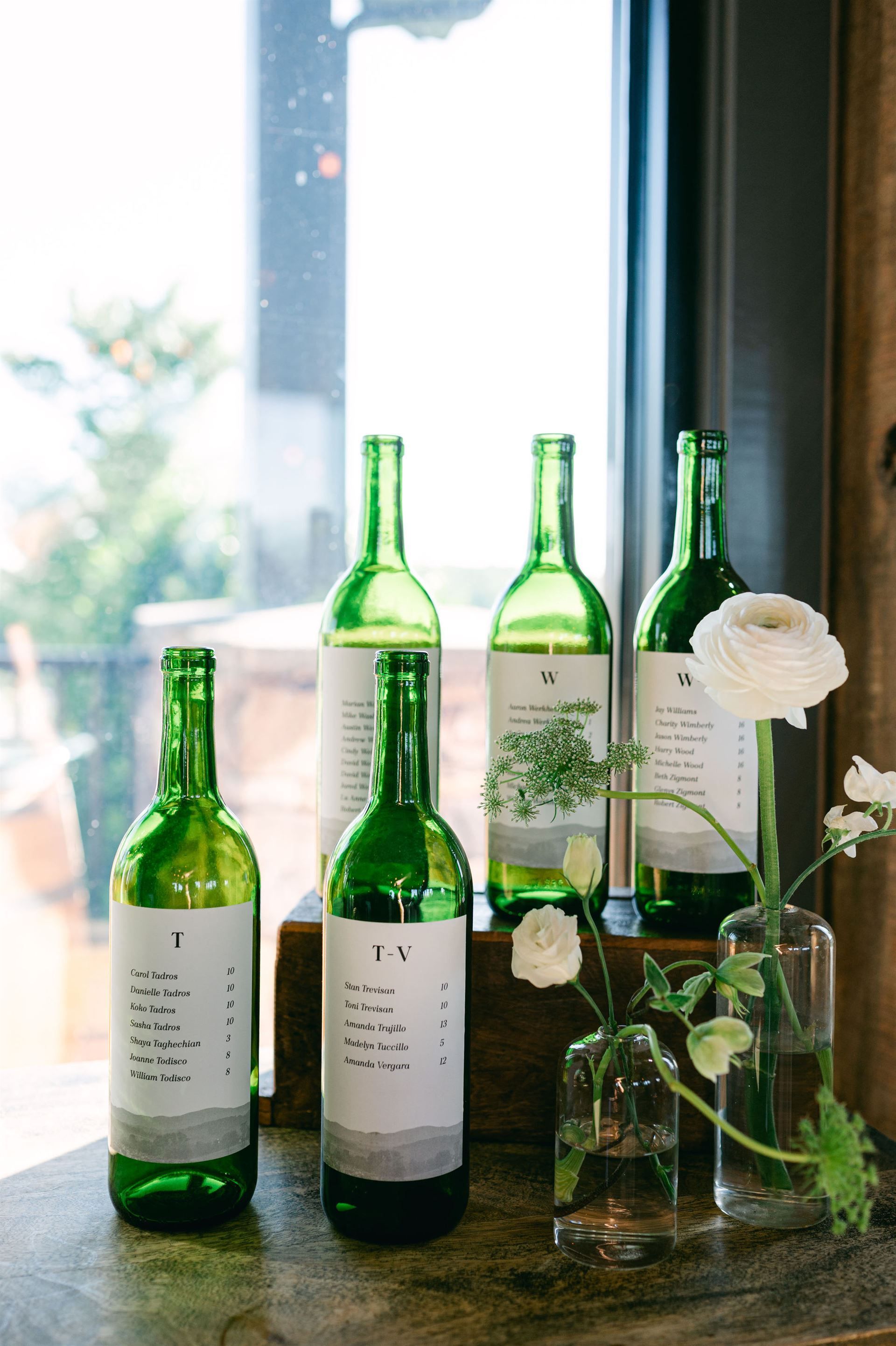 Green wine bottles with white labels, floral arrangements sit on a wooden surface by a window.