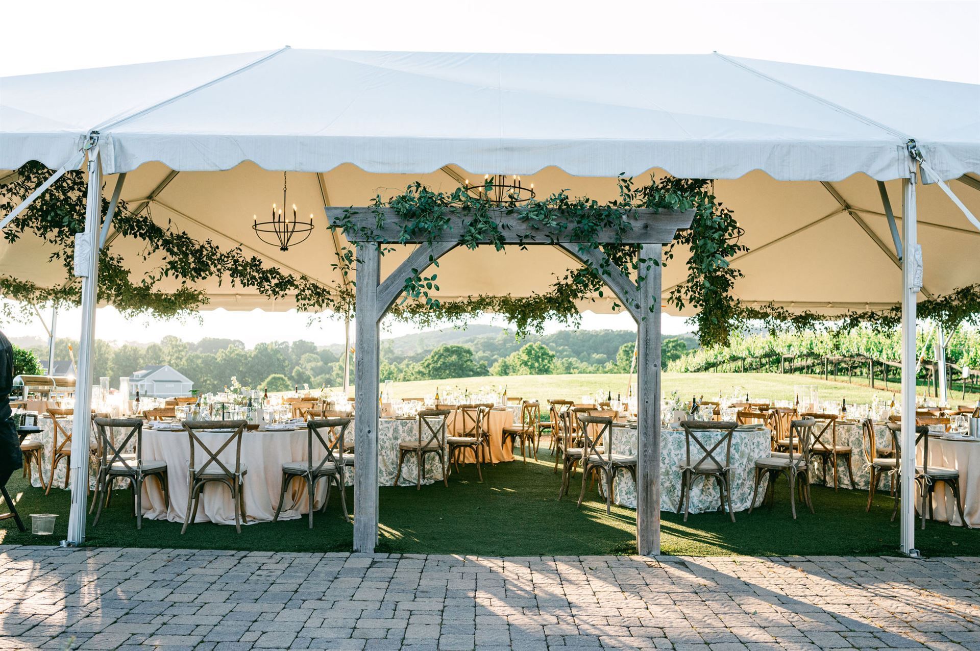 Wedding reception under a white tent with wooden arch decorated with greenery; tables and chairs set up outside.