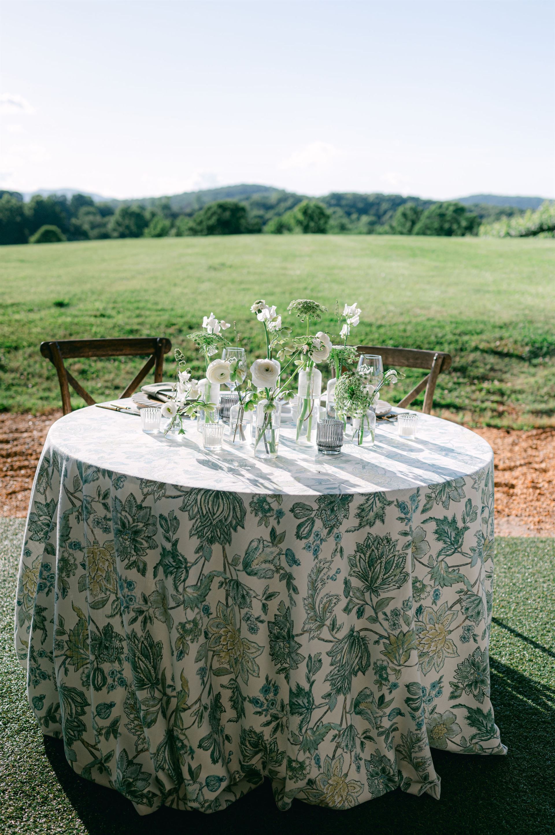 Round table set for an outdoor meal with floral tablecloth and flower vases. Green field and mountains in background.