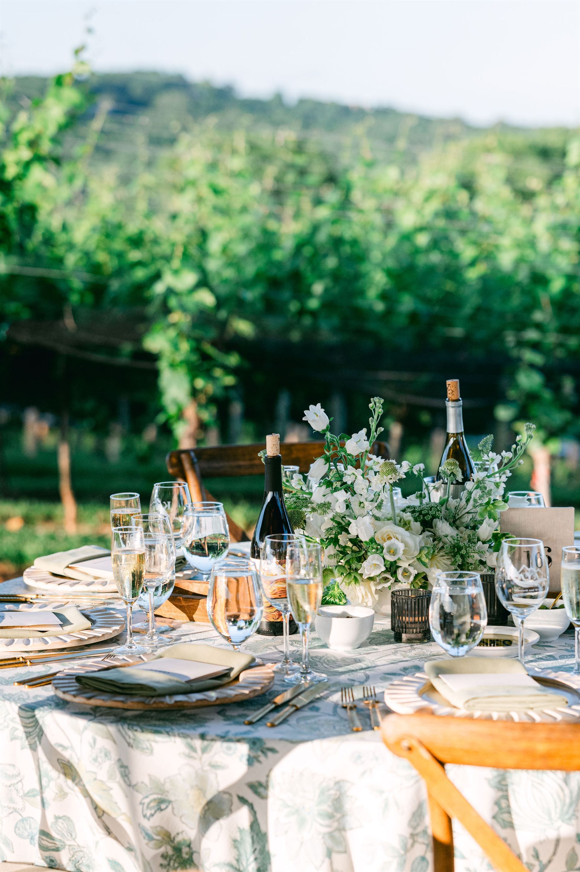 Elegant outdoor table setting with flowers, wine, and glasses in a vineyard.