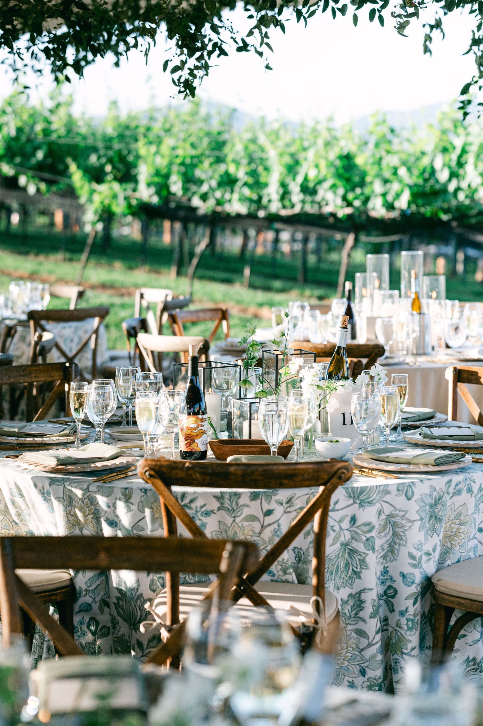 Wedding reception tables set in a vineyard, with floral tablecloths and wooden chairs.