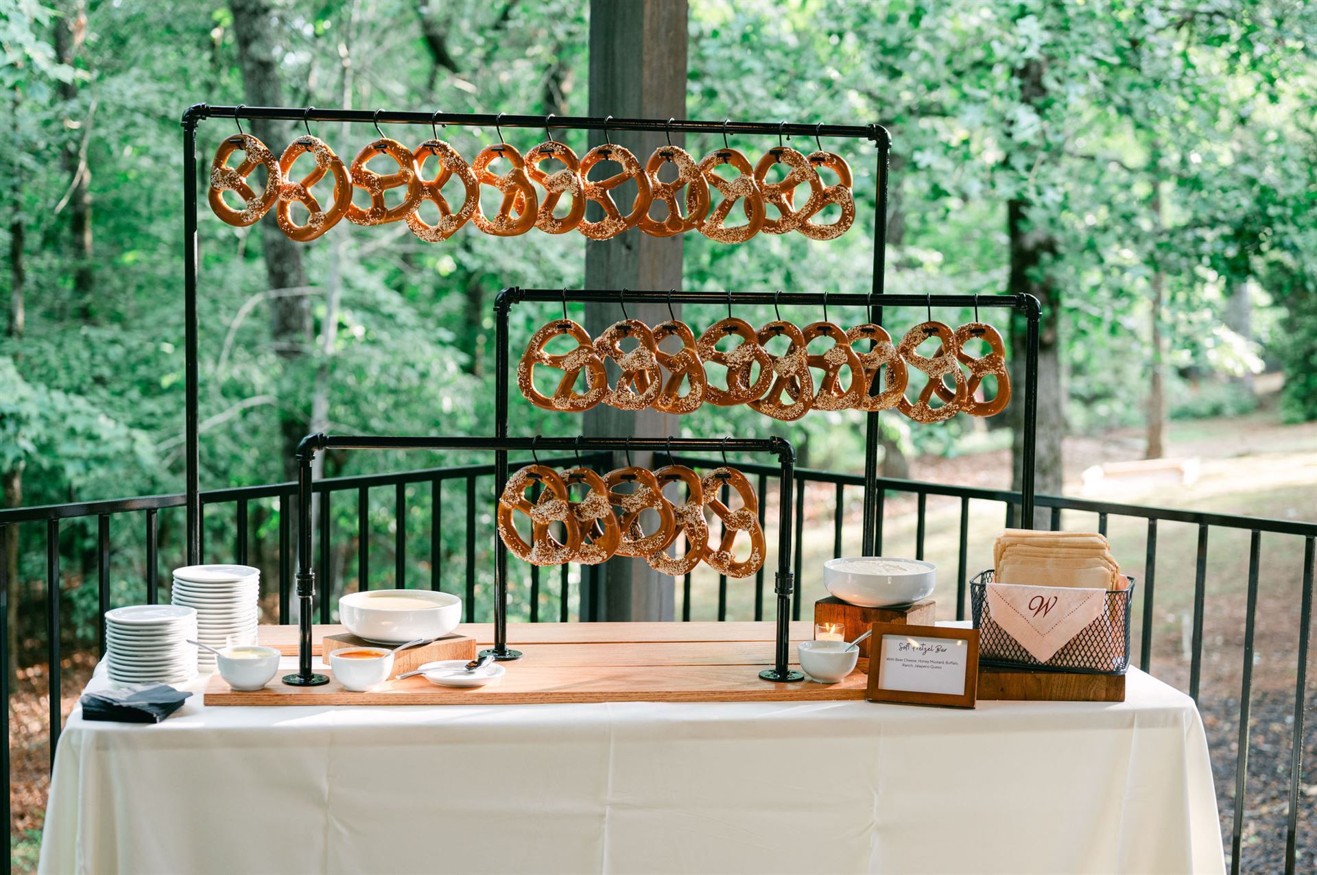 Pretzels hanging on a black metal display, set on a table with dips, outdoors with trees in the background.