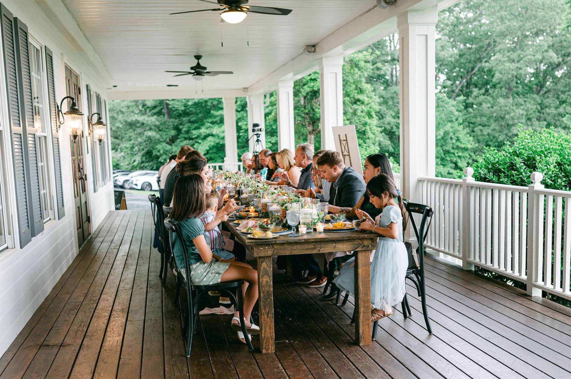 Long wooden table set for a meal on a porch, guests seated, surrounded by greenery.