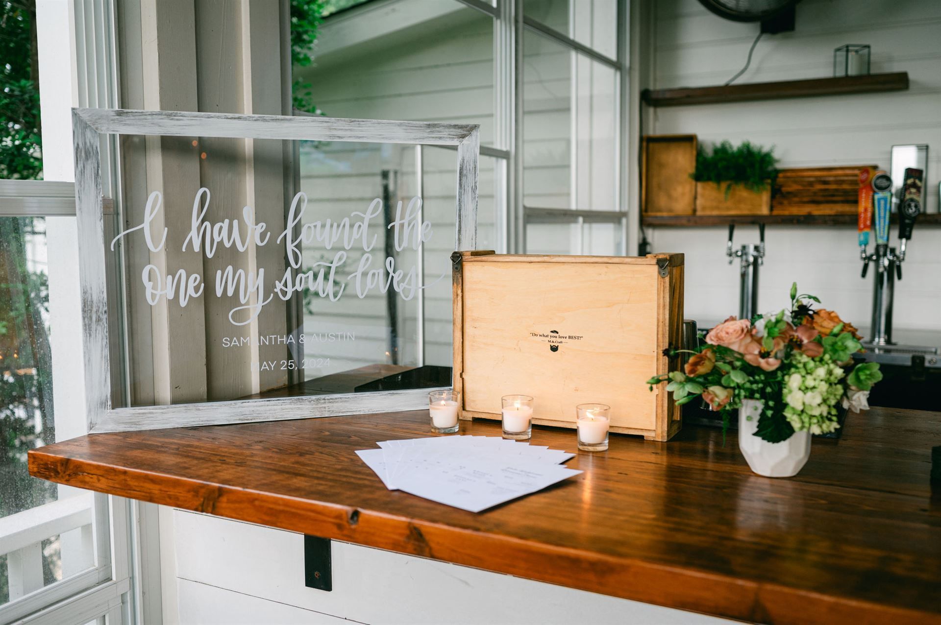 Wedding reception table with sign, box, candles, flowers, and guest book, next to a bar.