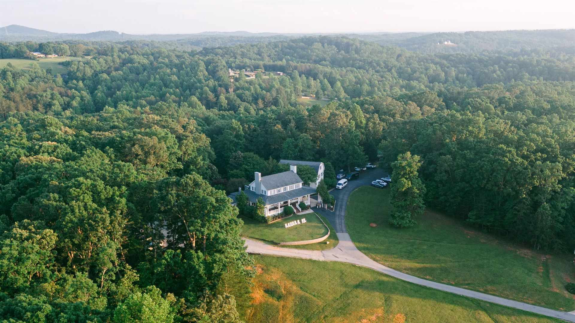 Aerial view of a house nestled in a lush green forest, with a winding driveway and clear sky.