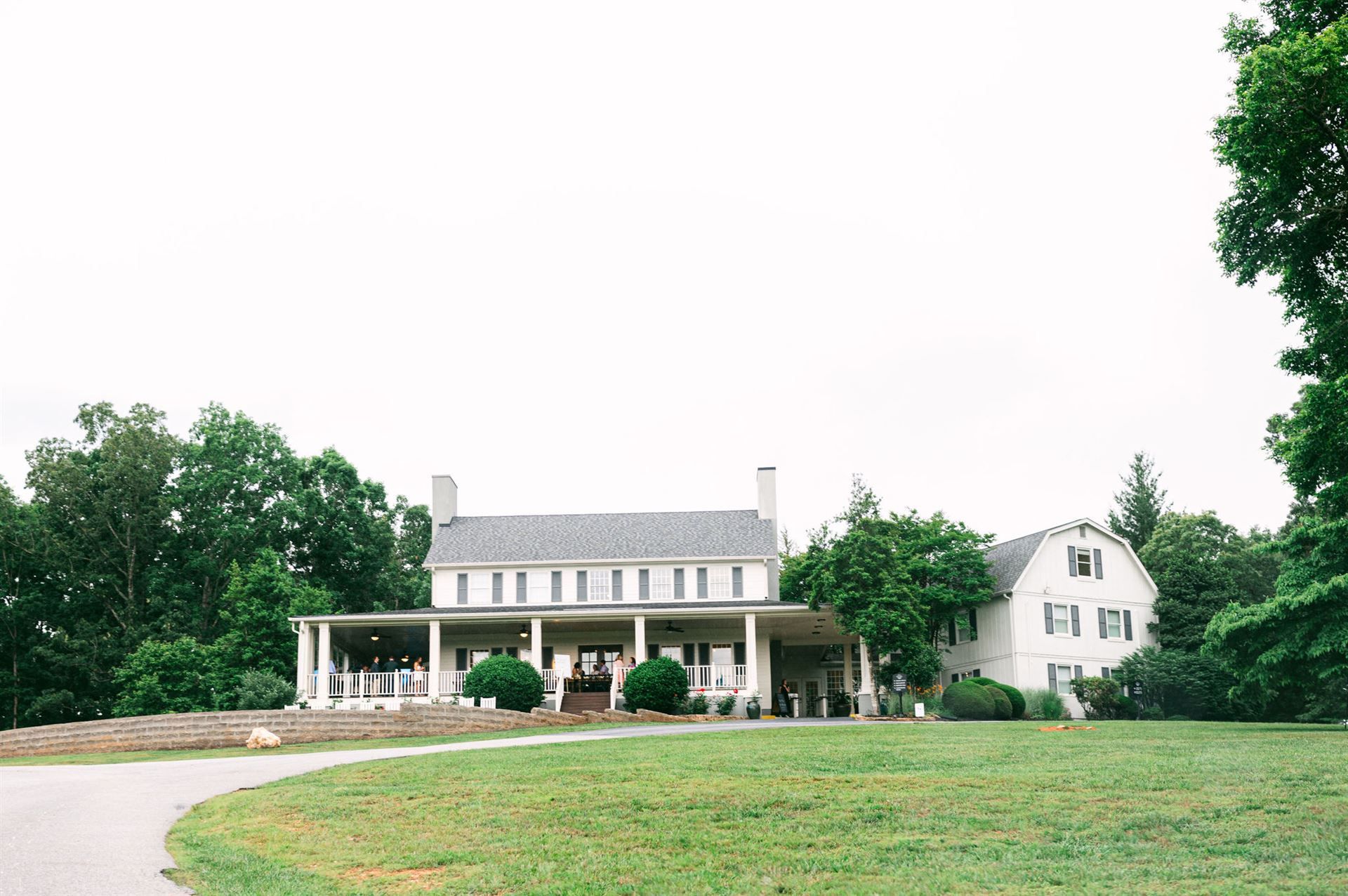 White two-story house with wrap-around porch, gray roof, and green lawn, trees surround the building.