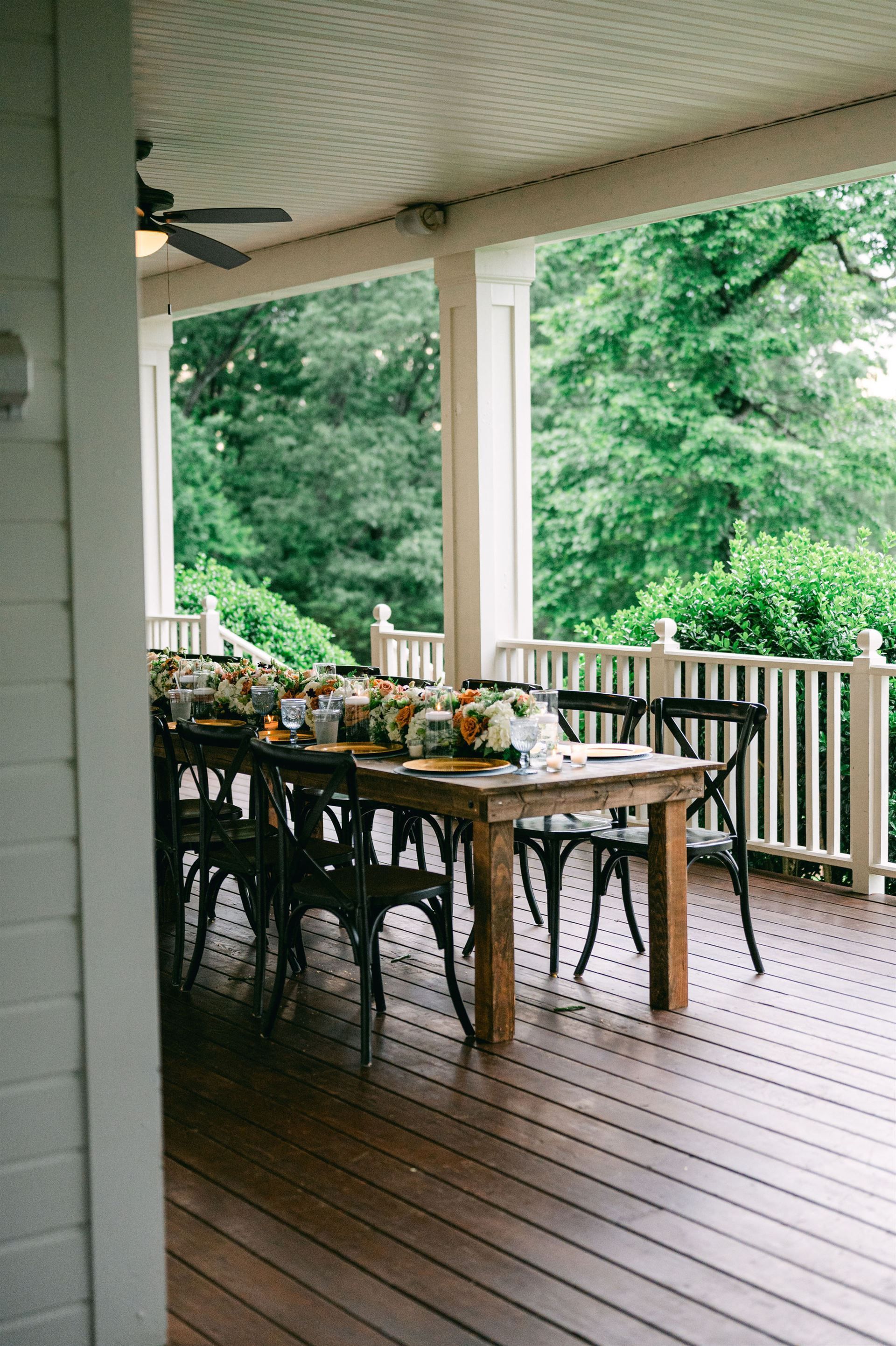 Wooden table set for a meal on a porch, surrounded by chairs. Lush green trees are visible in the background.