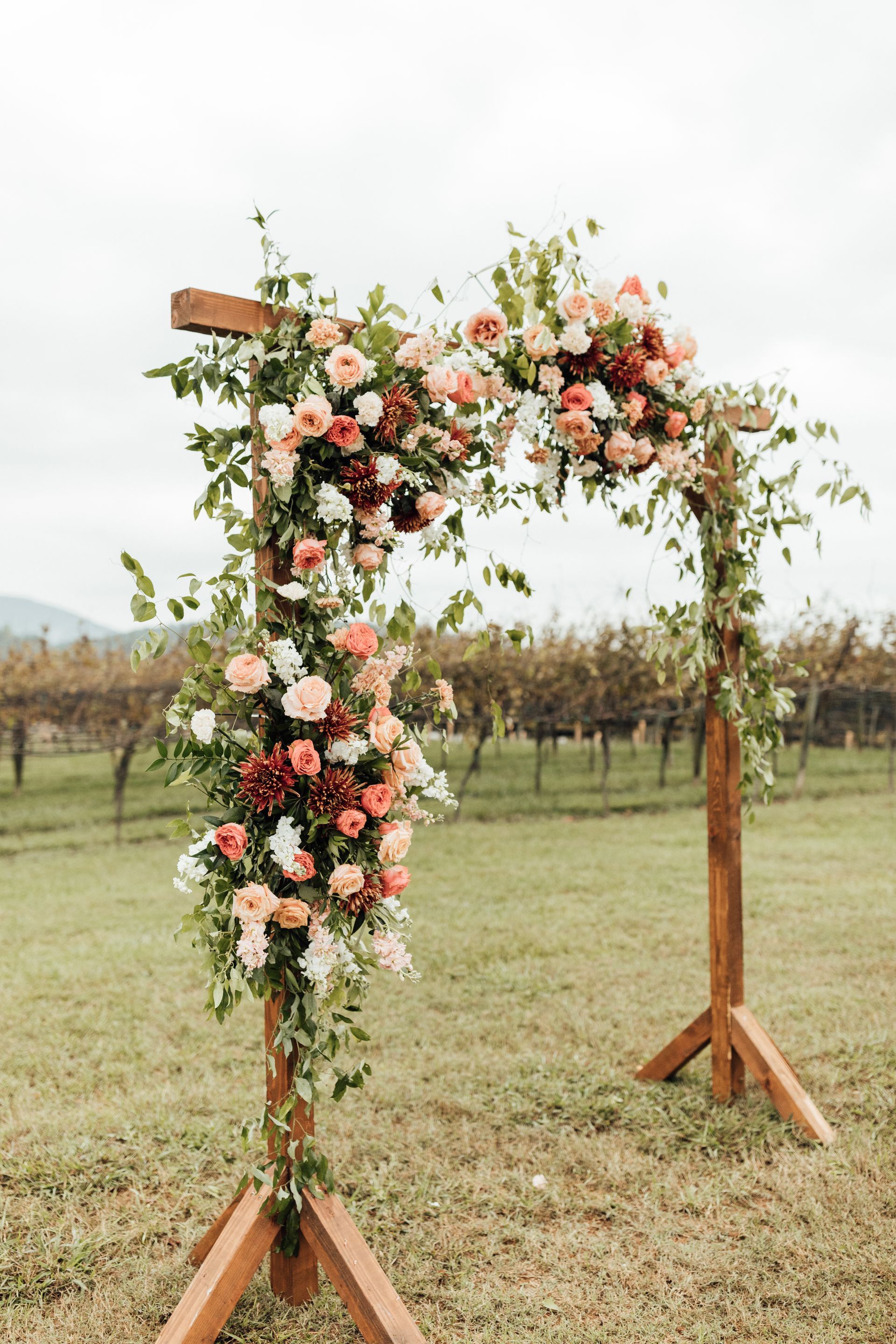 Wooden wedding arch decorated with pink and white flowers, vines, and set in a field.