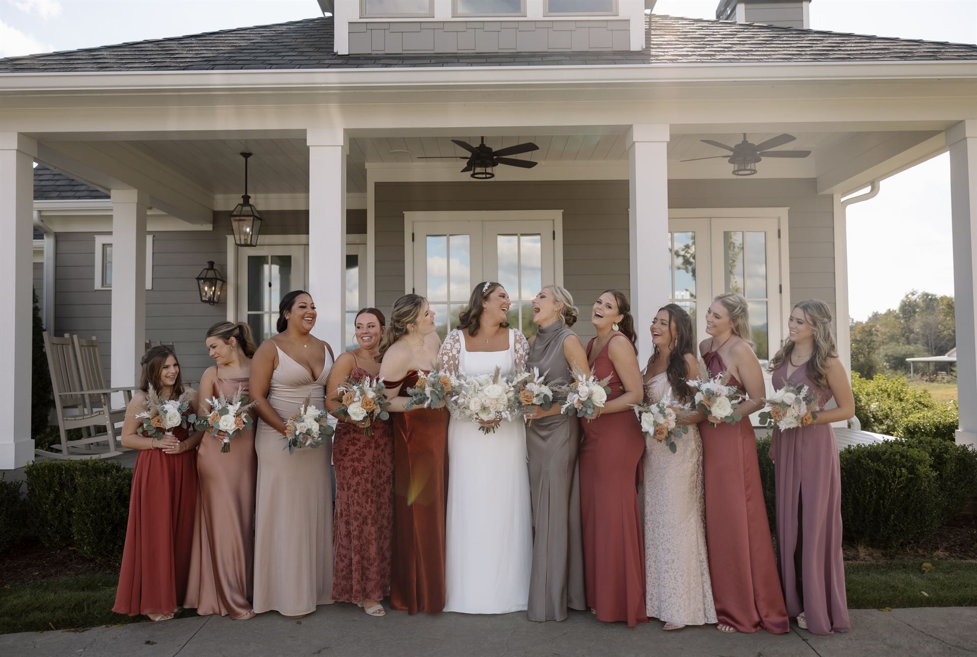 Bride and bridesmaids laugh in front of a house, wearing different colored dresses and holding bouquets.