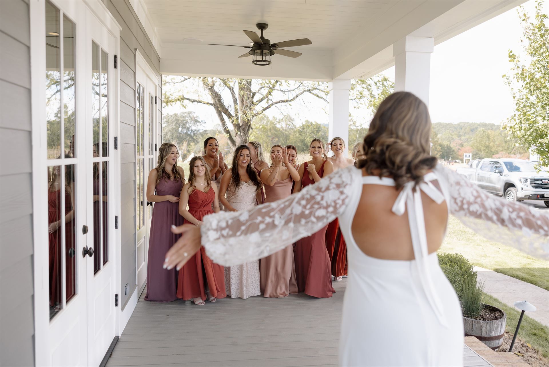 Bride in white dress, arms outstretched, facing bridesmaids on a porch; they react with surprise and delight.