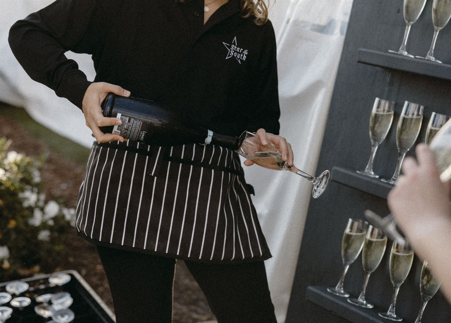 Server pours champagne into a glass from a bottle, wearing a black shirt, striped apron.