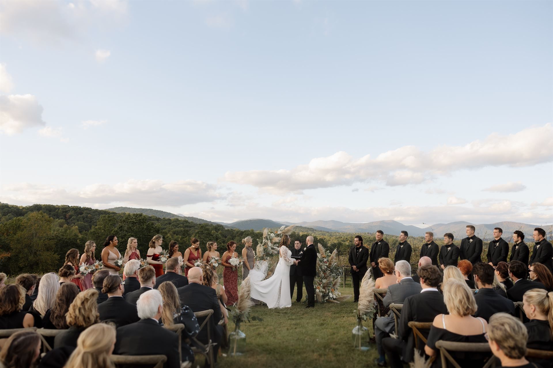 Wedding ceremony outdoors; couple under arch, wedding party and guests seated, mountains in background.