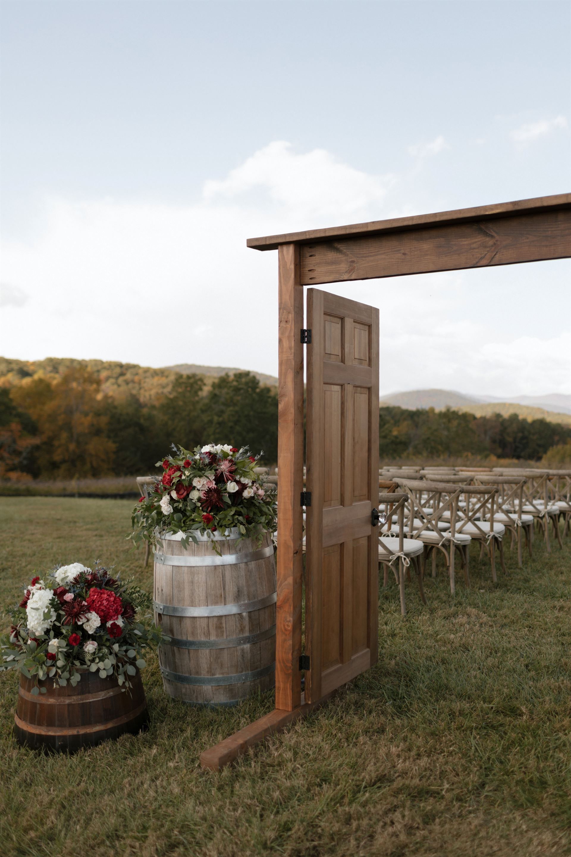 Wedding ceremony setup with wooden door, barrels of flowers, and chairs in a field with a mountain backdrop.
