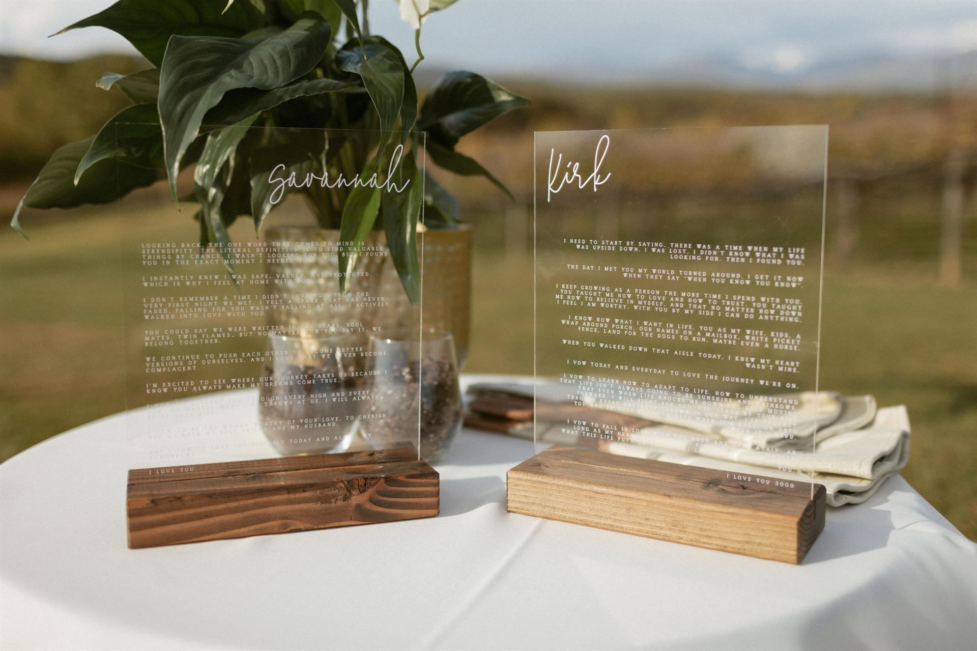 Clear acrylic wedding signs on wood blocks on a white tablecloth, with greenery in the background.