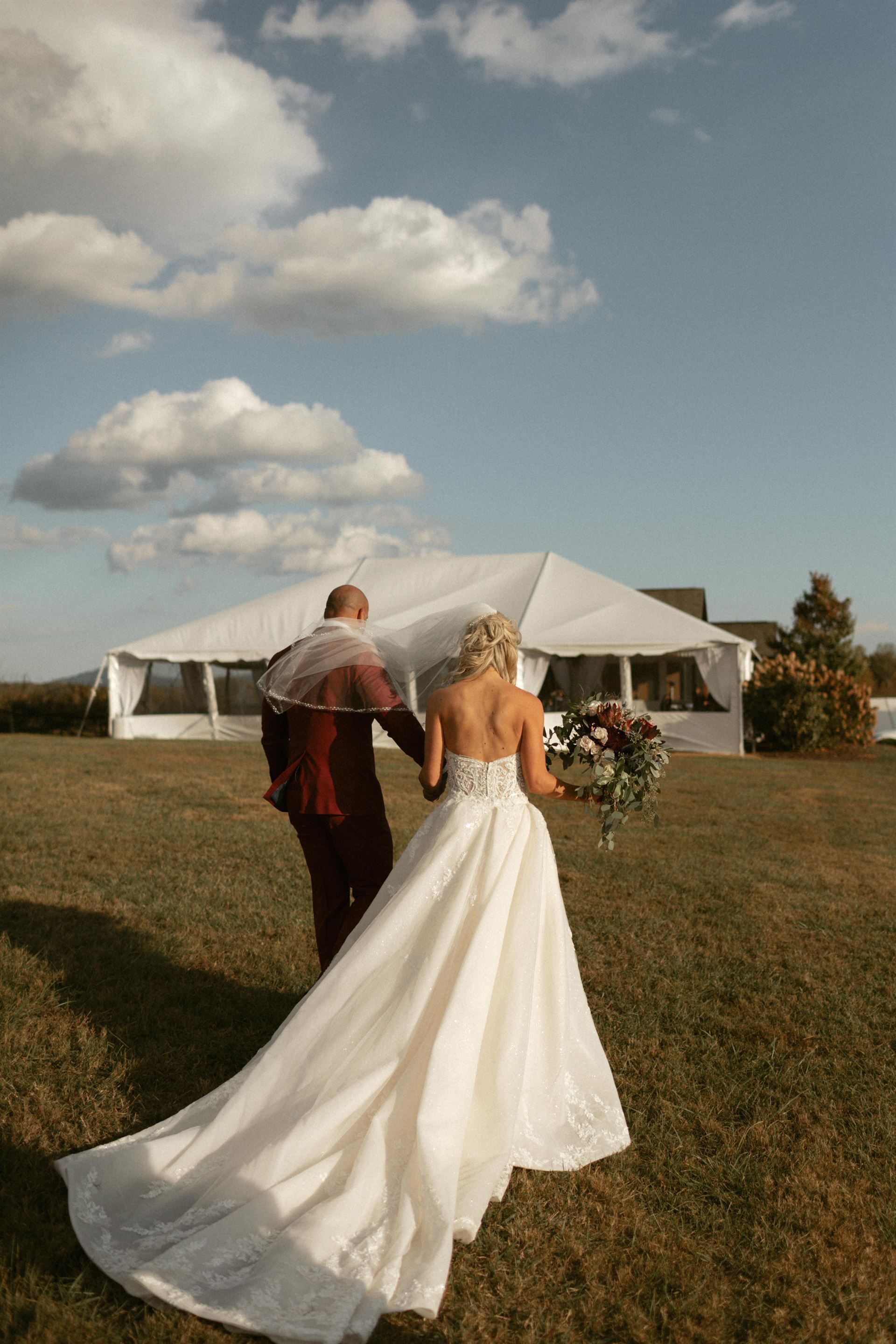 Bride in white gown and groom in burgundy suit walk toward a tent on a sunny day.