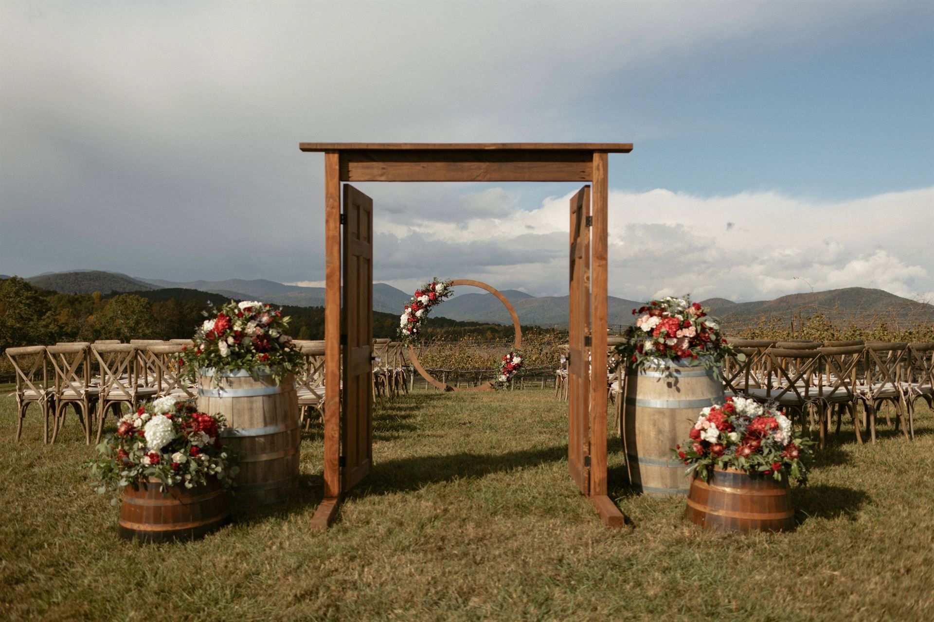 Wedding ceremony setup outdoors with floral barrels, wooden arch, and vineyard backdrop.