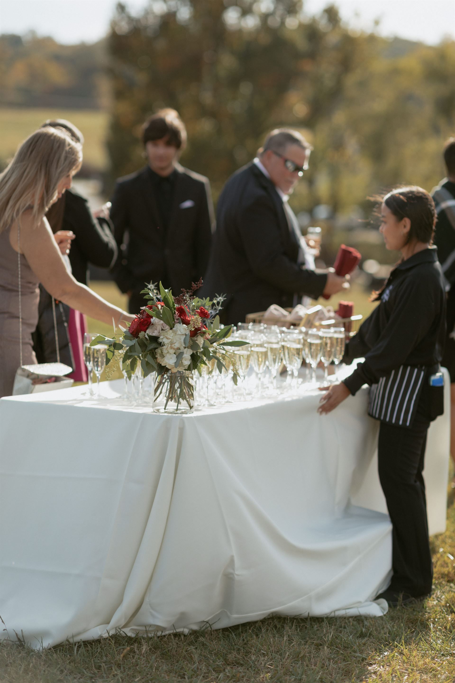People at a table with champagne flutes and flowers outdoors. A server refills a glass.