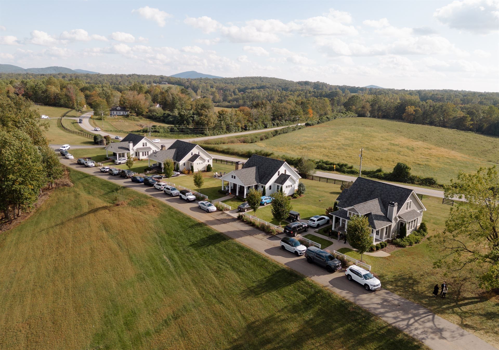 Aerial view of homes with a line of cars on a gravel road, fields, trees, and rolling hills in the background.