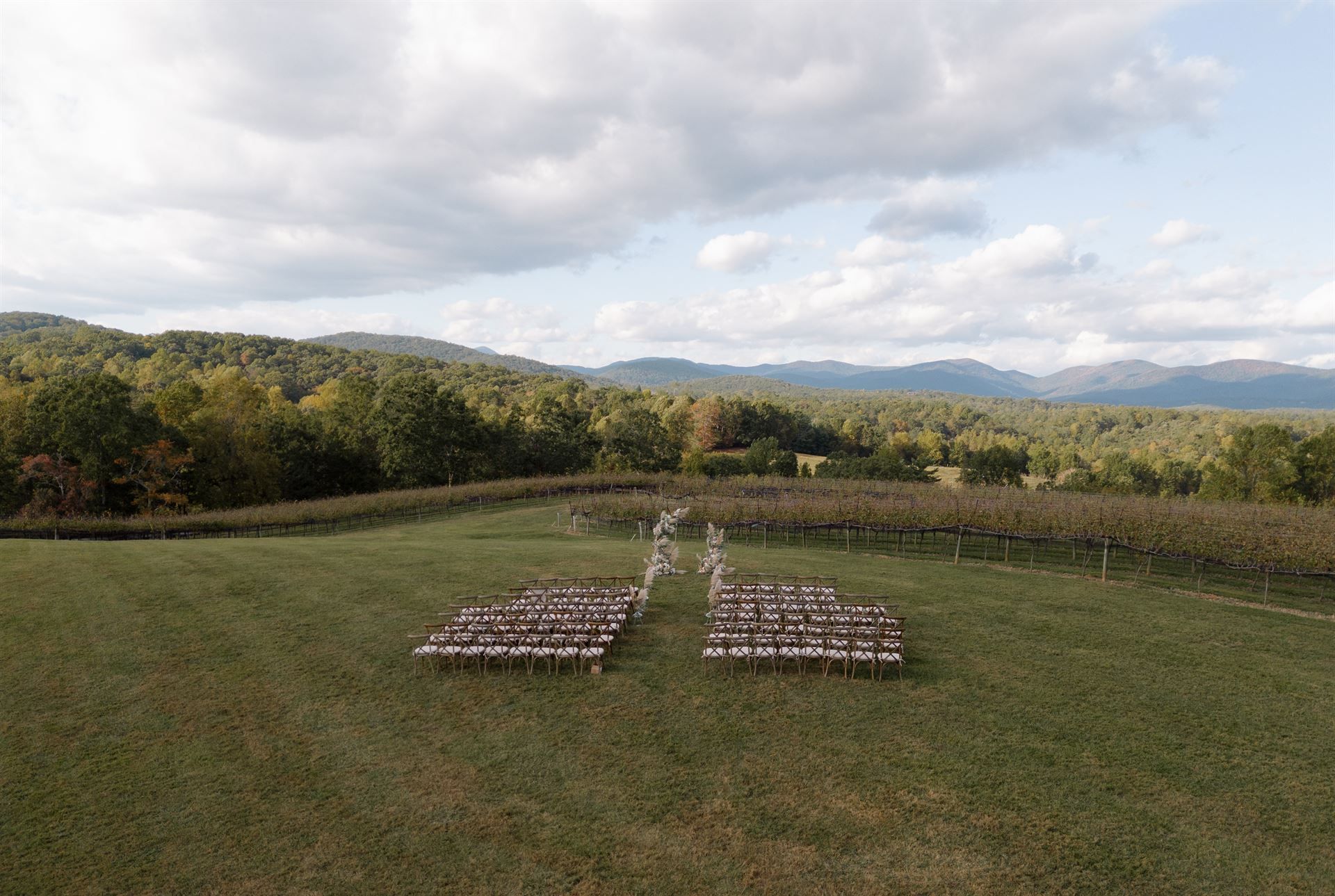 Wedding ceremony setup in field with rows of chairs, archway, and trees under a cloudy sky.