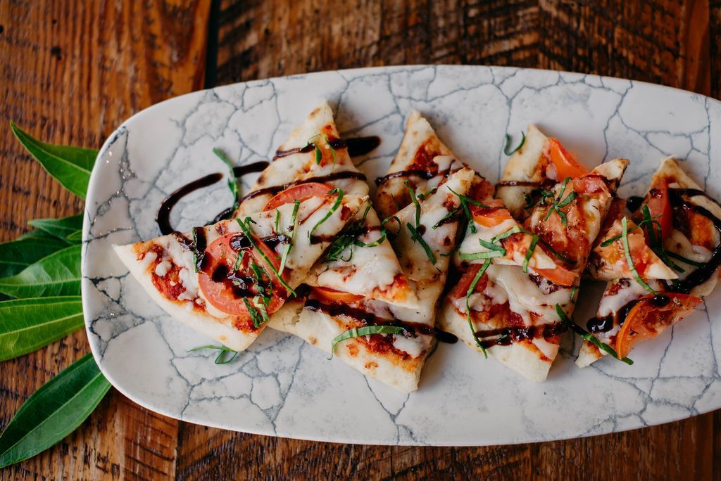 Flatbread slices with cheese, tomato, herbs, and balsamic glaze on a decorative plate, set on a wooden table.