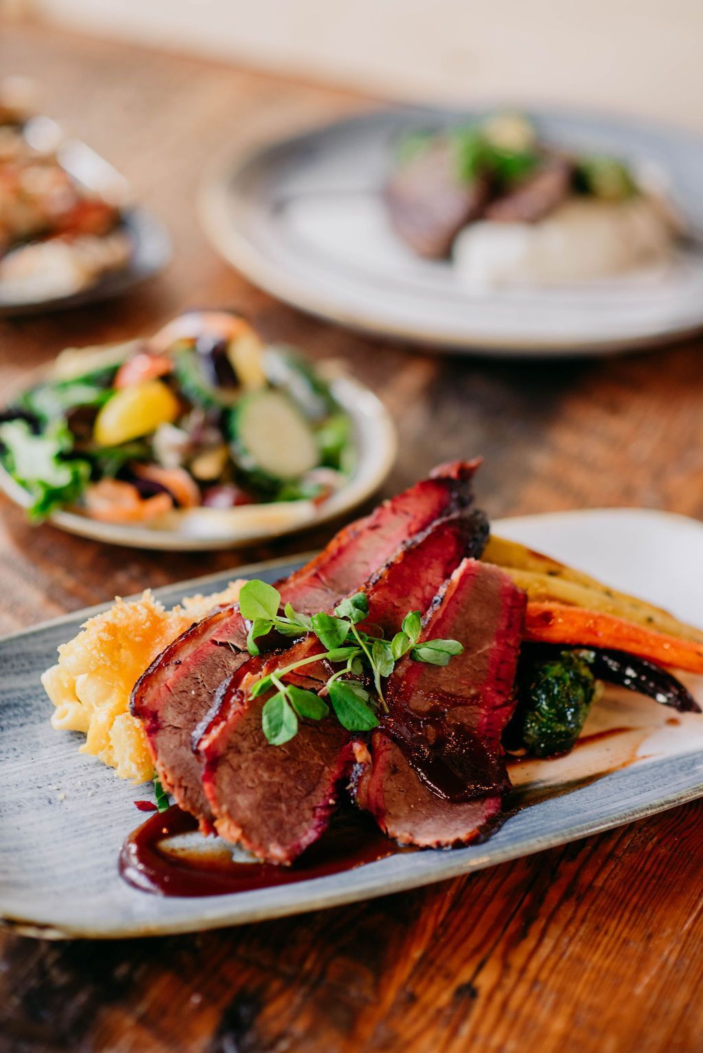 Sliced meat with gravy, mashed potatoes, and roasted vegetables on a plate, set on a wooden table.