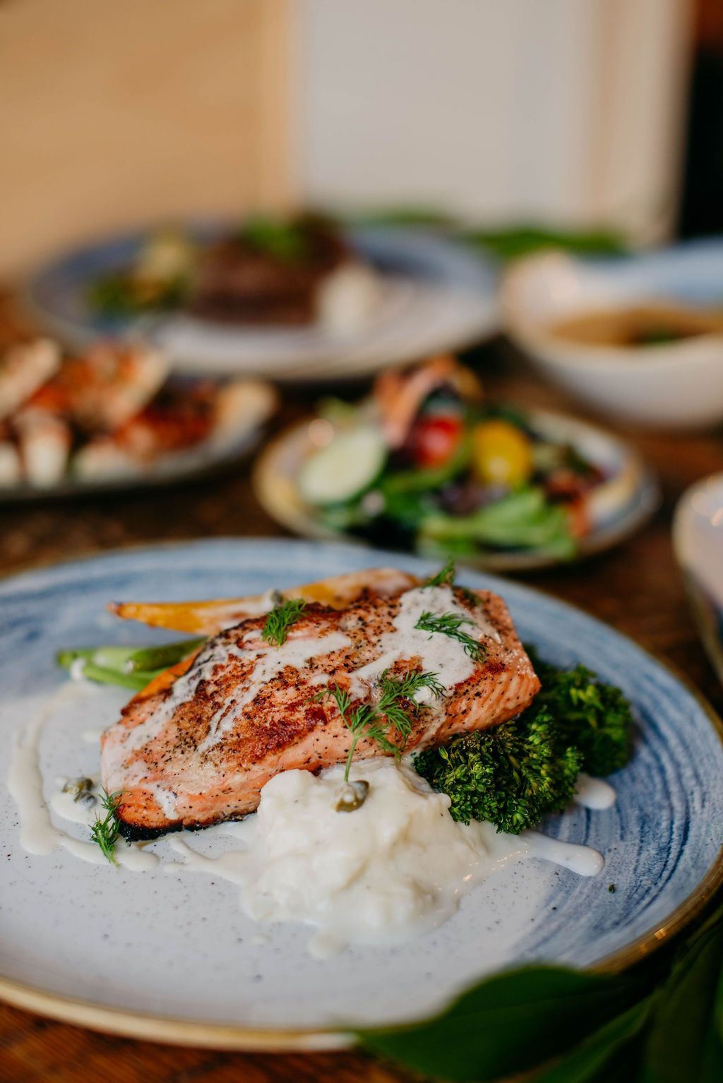Grilled salmon with sauce, broccoli, and vegetables on a blue-rimmed plate. Other dishes visible.