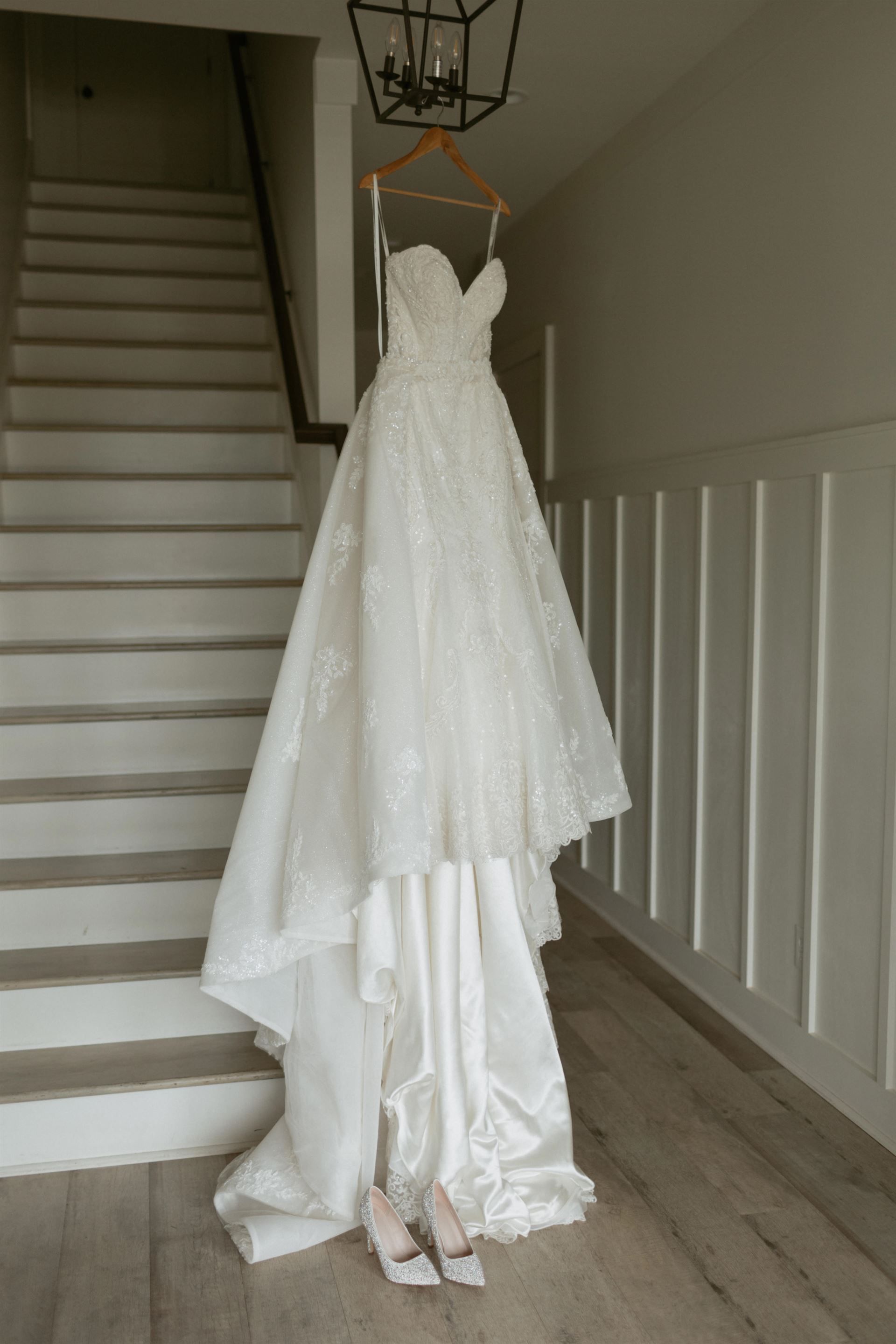 Wedding dress hanging on a wooden hanger by a staircase. White dress with long train. Beige heels on the floor.