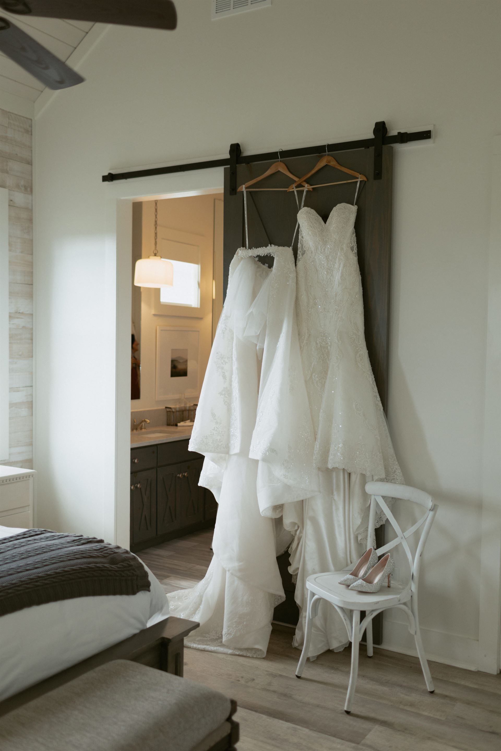 Two white wedding dresses hanging on a sliding barn door; a white chair and shoes nearby.