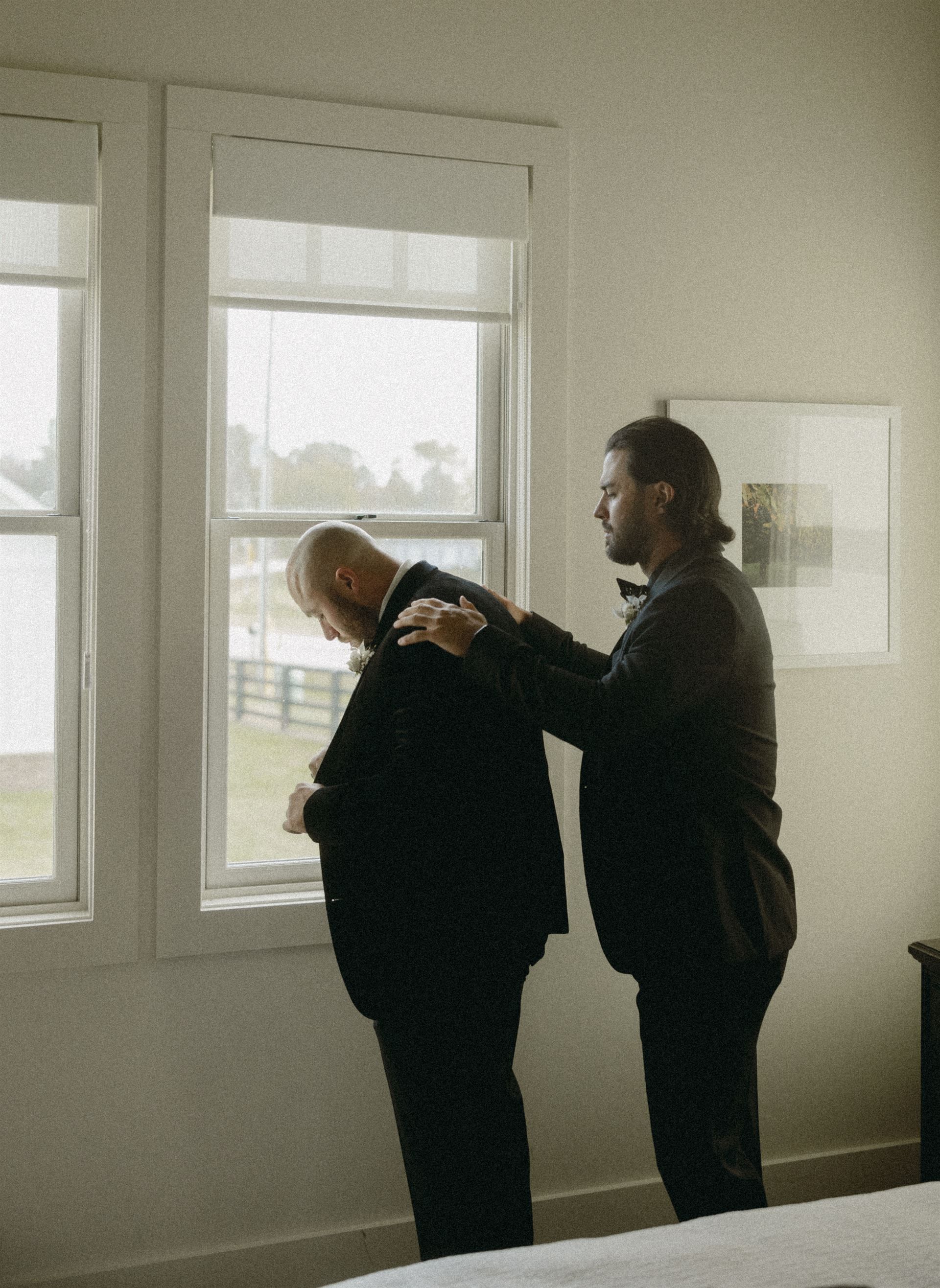 Man helps another button a suit jacket by a window.