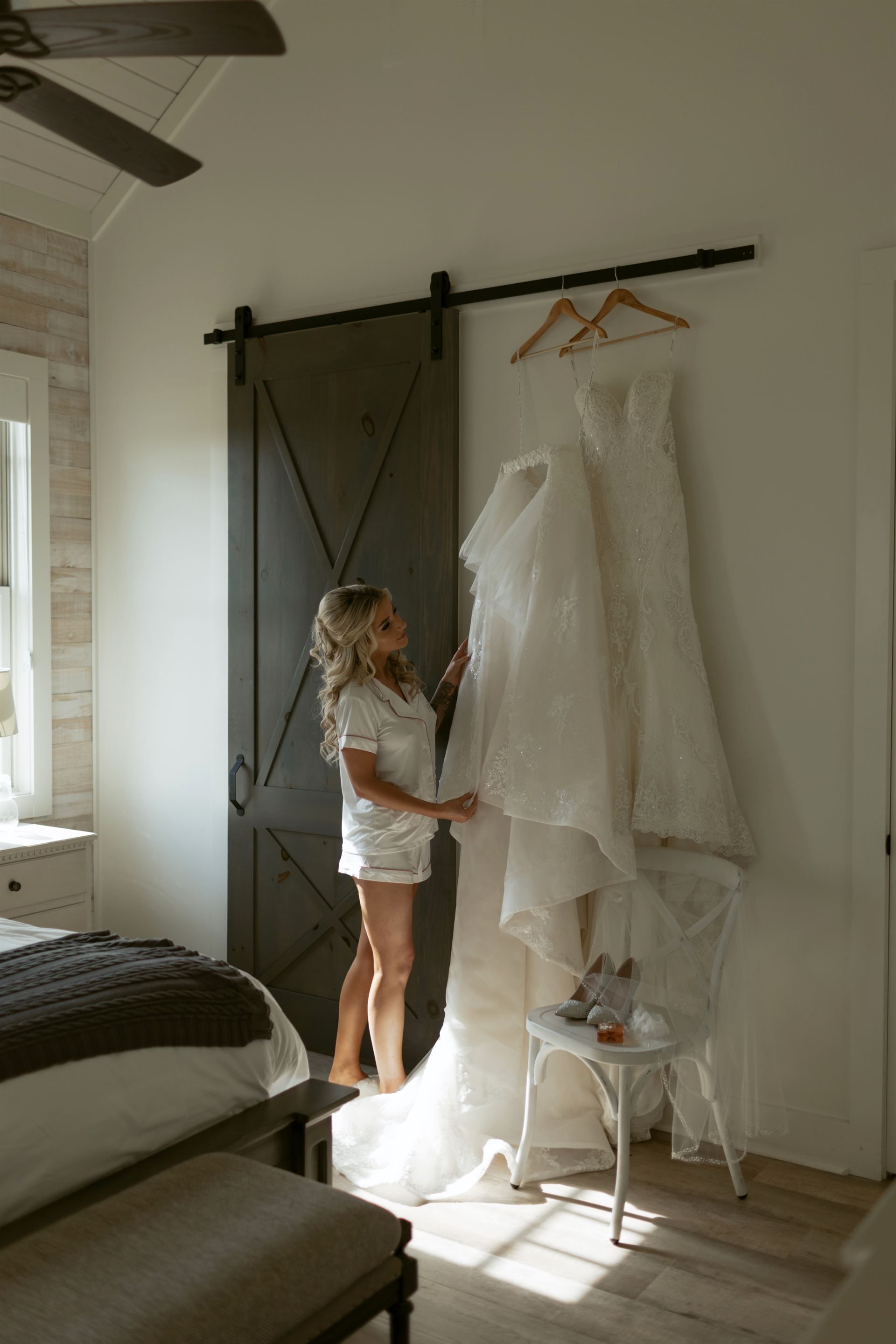 Woman admires wedding dresses hanging on a rail near a barn door in a bedroom, with a bed in the foreground.