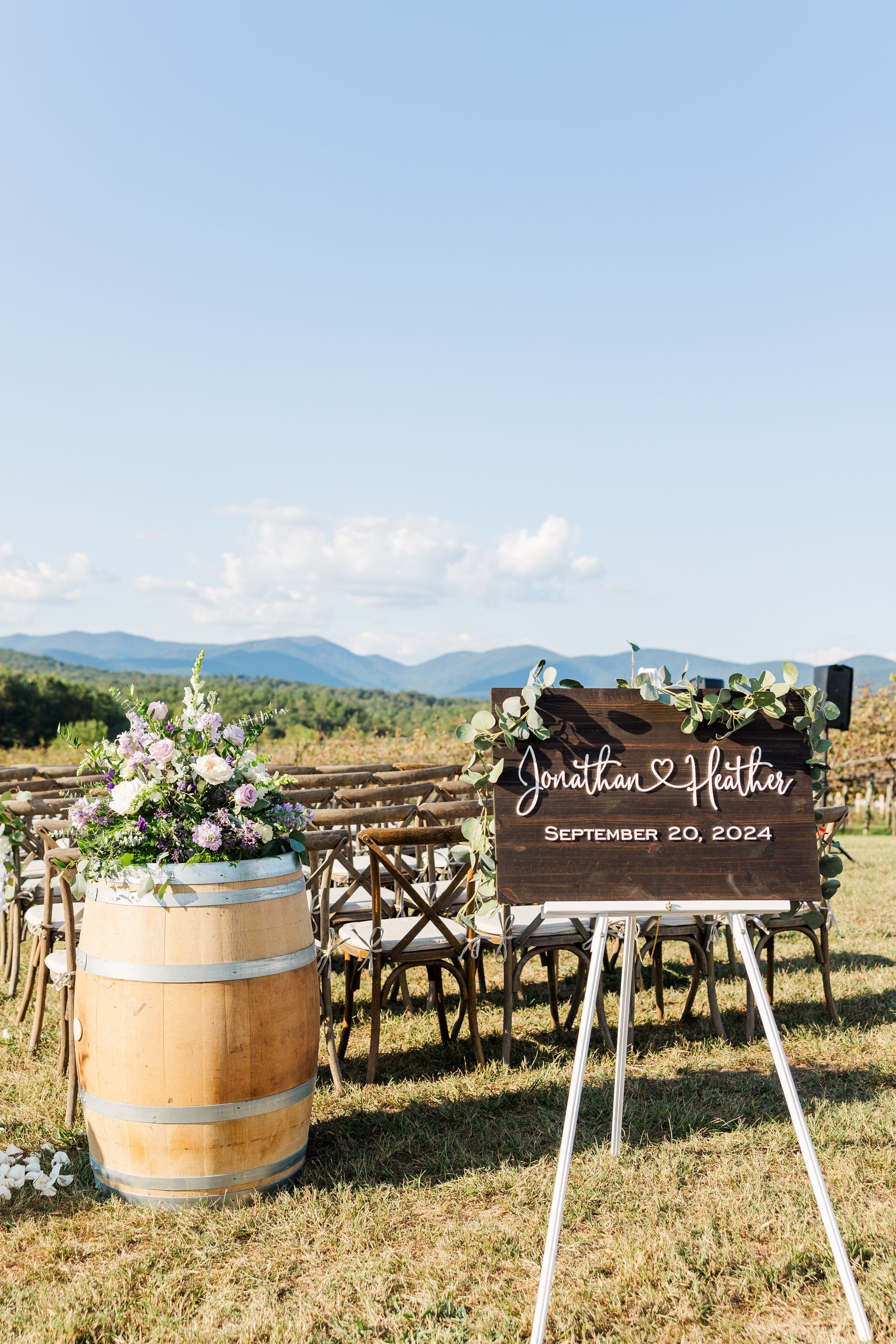 Wedding ceremony setup with a sign and barrel decorated with flowers. Wooden chairs face the mountains.