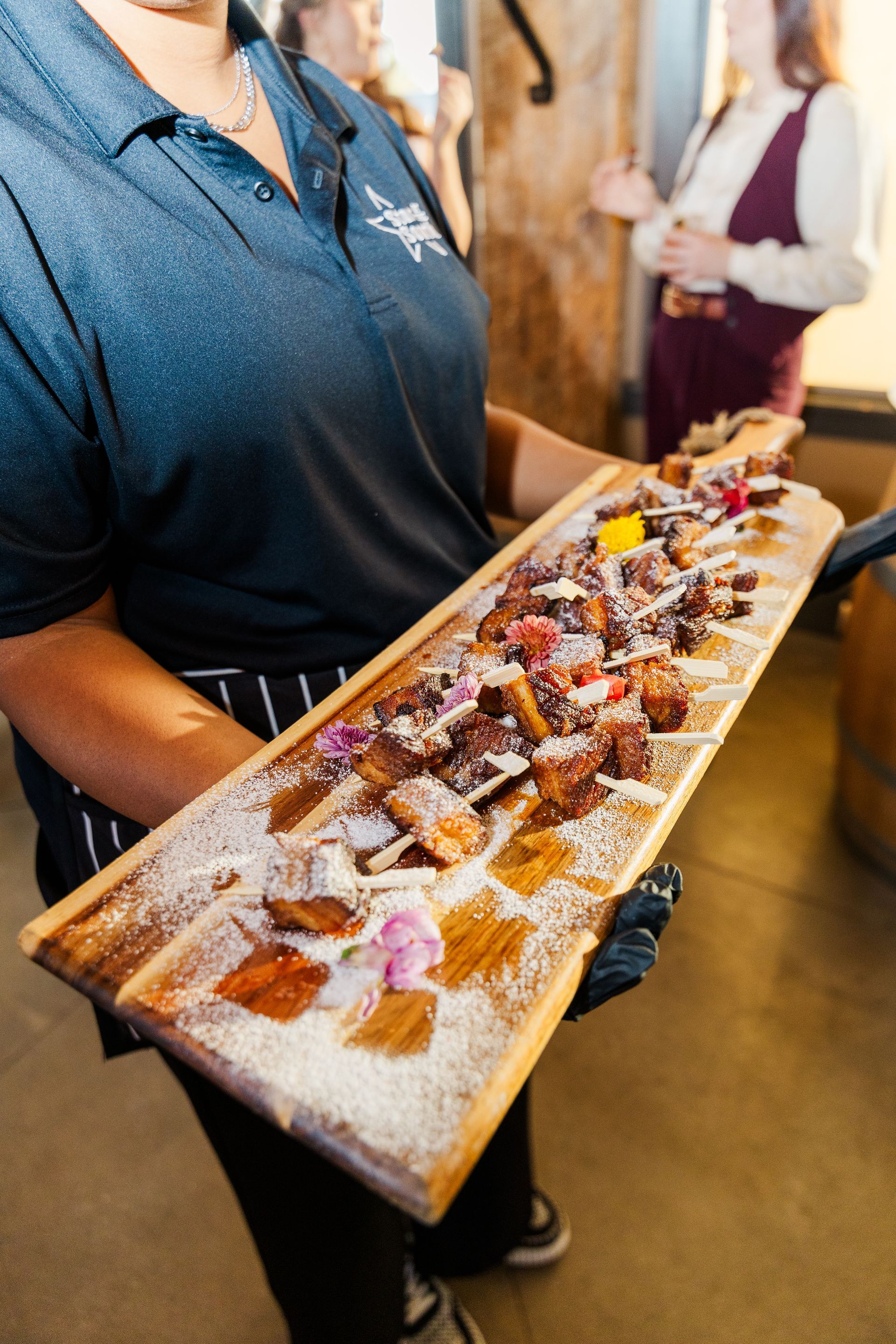 Server holding a long wooden tray of food skewers with powdered sugar and colorful garnishes.