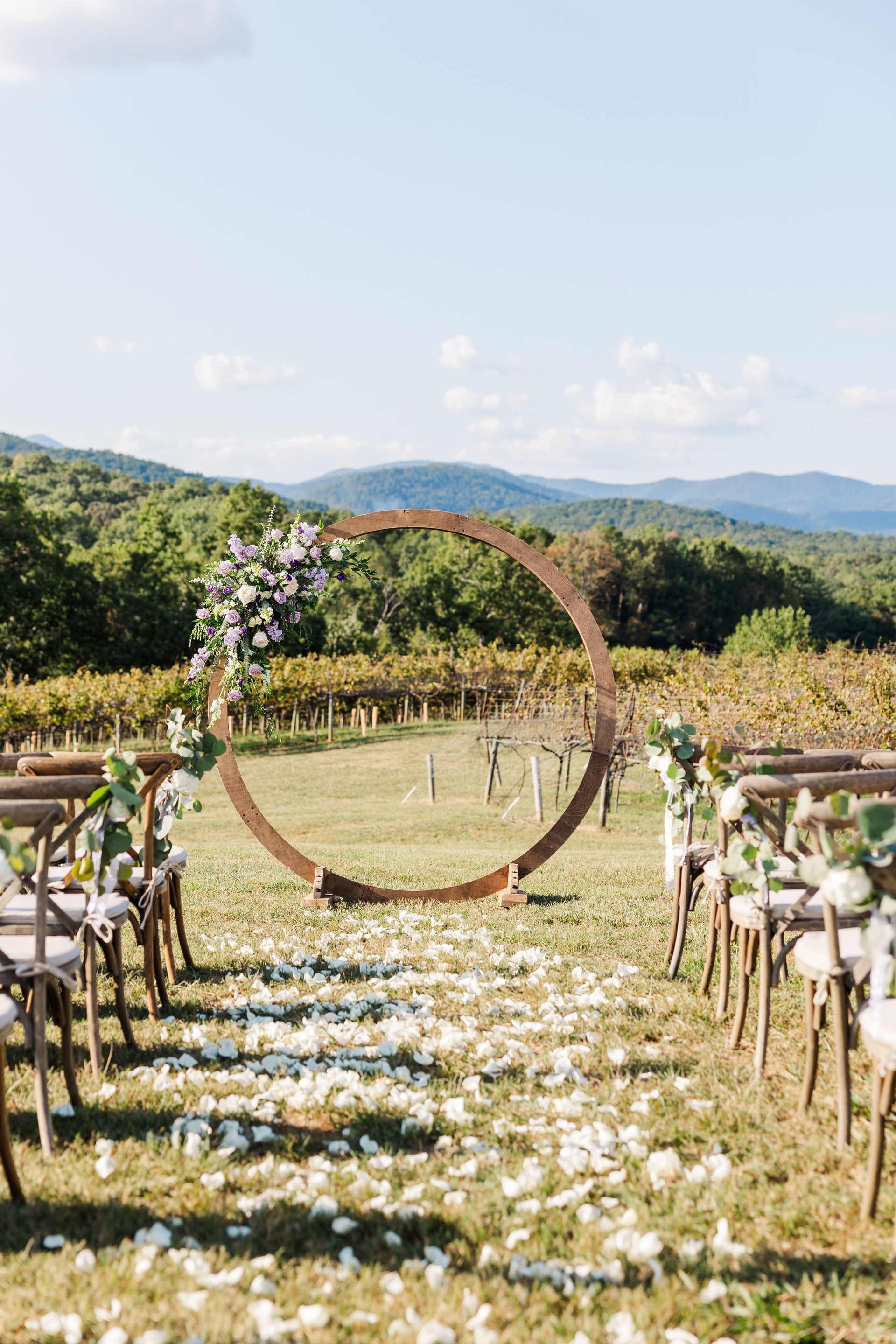 Wedding ceremony setup with round arch, vineyard, mountain backdrop. Chairs line aisle with petals.