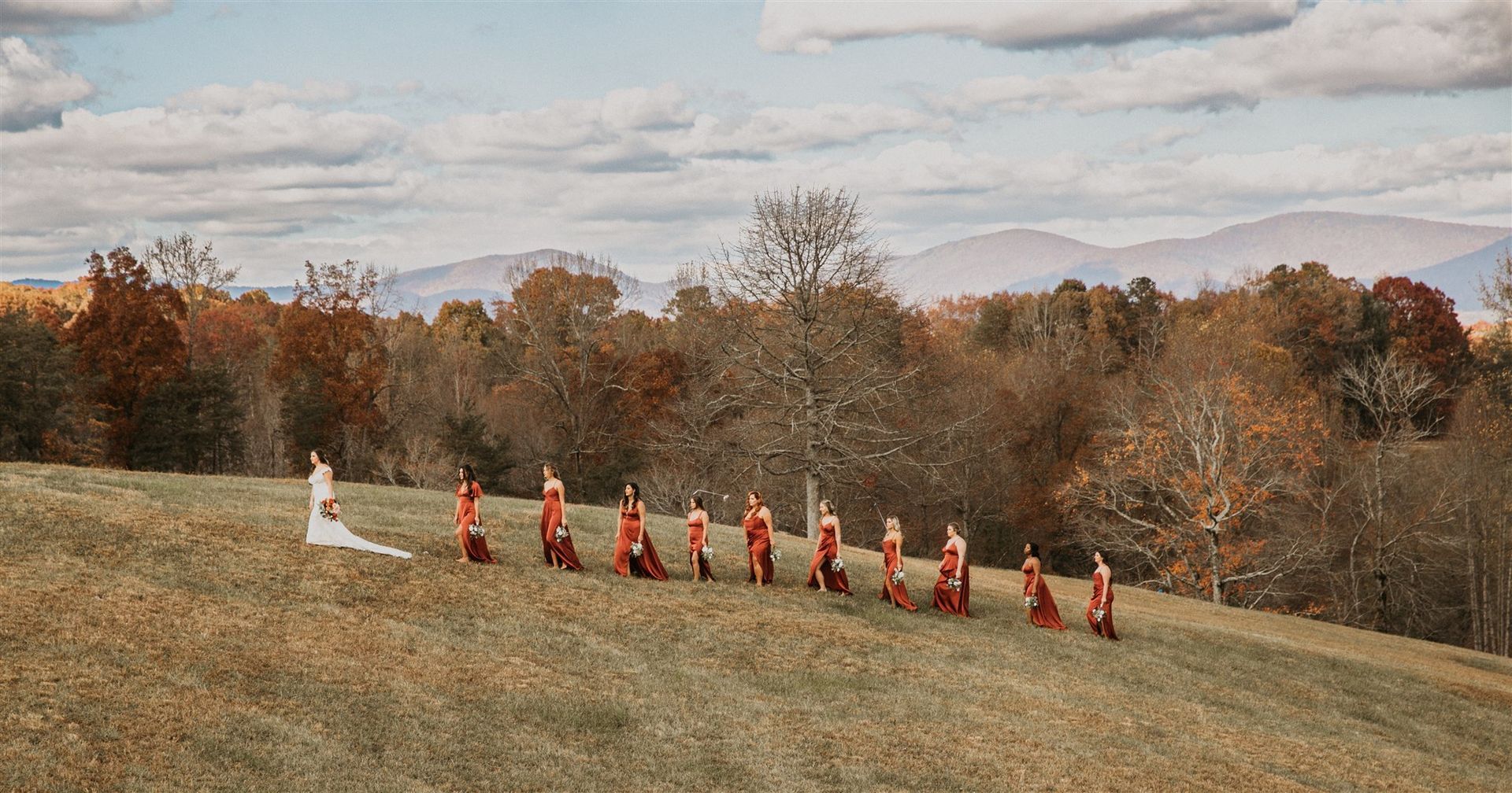 Bride in white dress with bridesmaids in red dresses, standing on a grassy hillside in fall. Mountains and trees in the background.