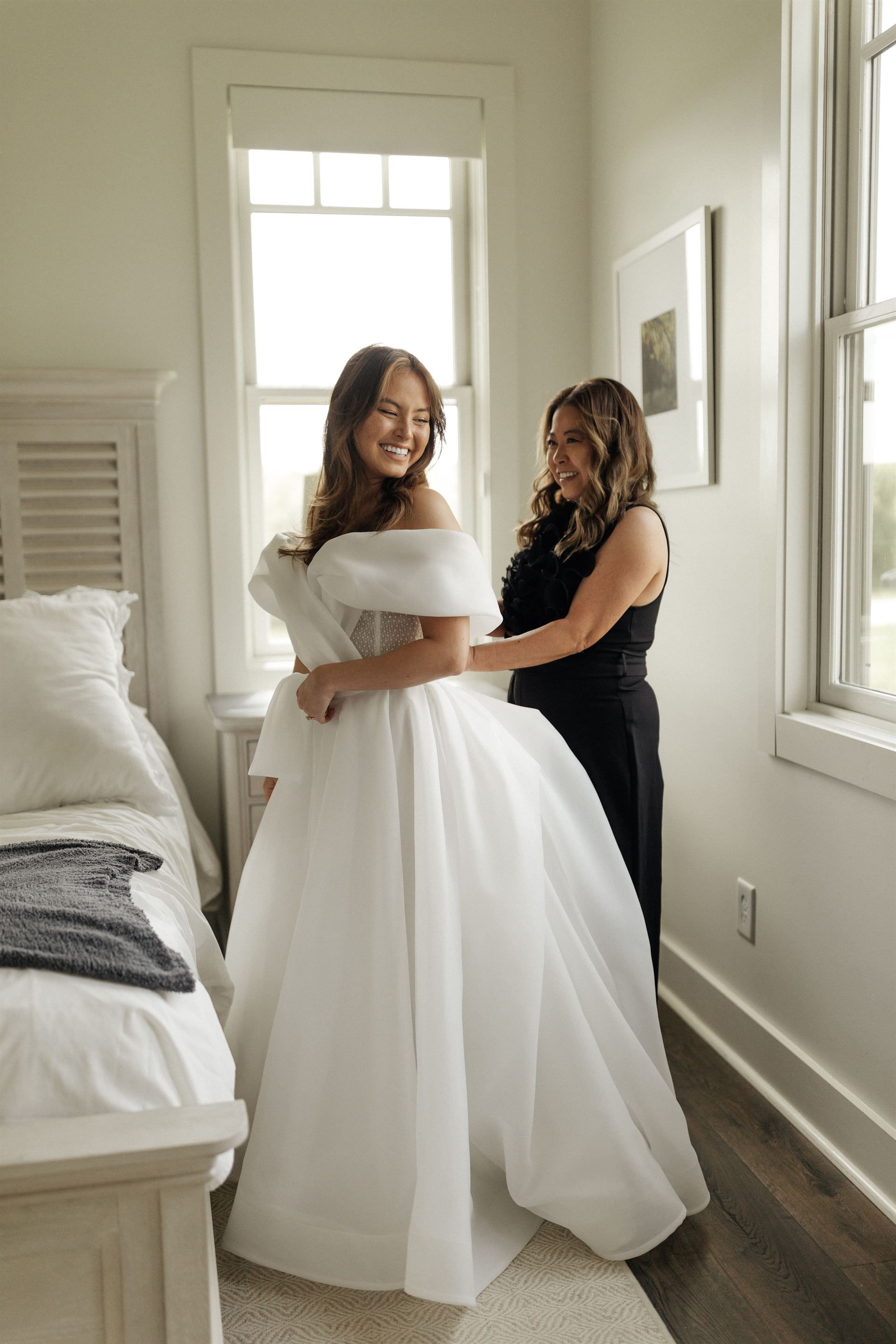 Woman in a white wedding dress smiles as another woman helps adjust the dress in a bedroom with a window.