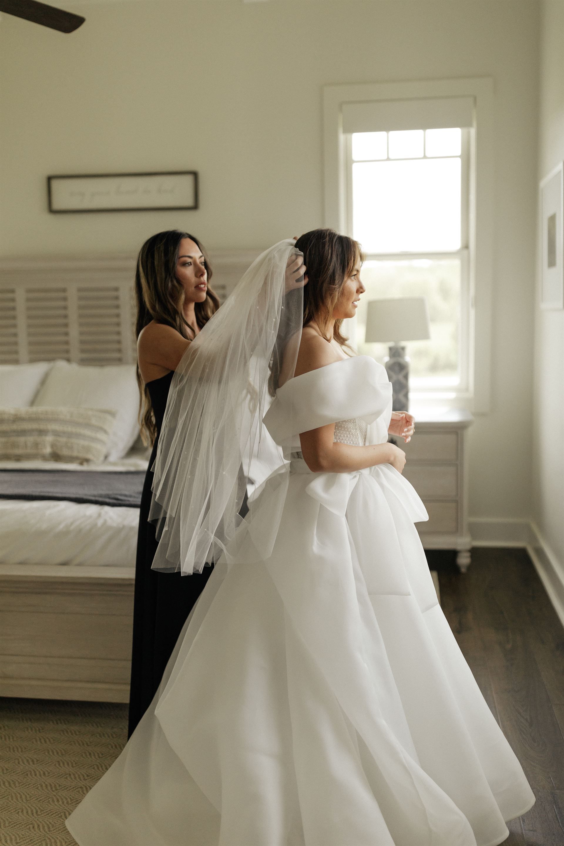 Woman in white wedding dress, veil being placed by another woman in black dress, bedroom setting.