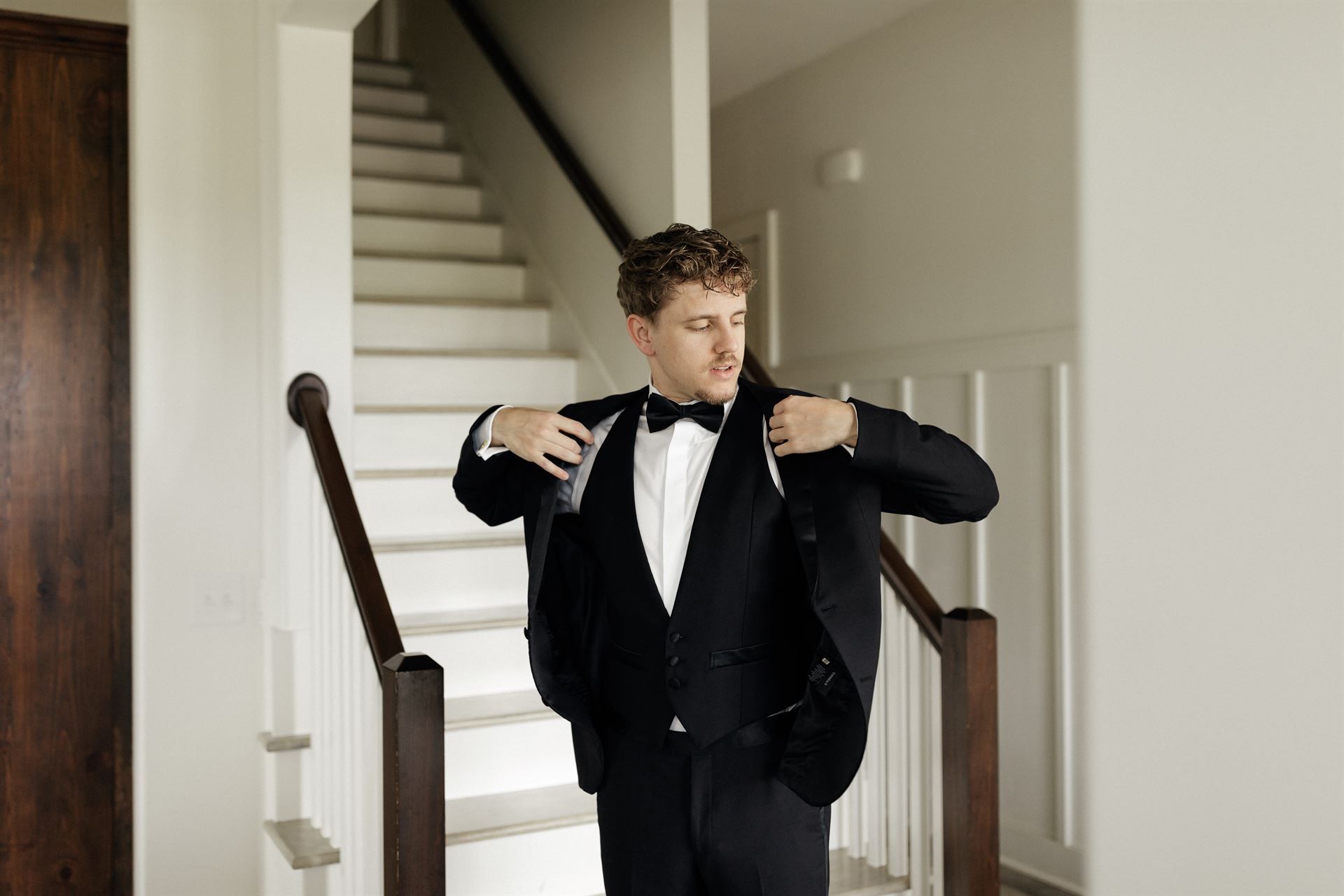 Man in tuxedo adjusting his jacket, standing on a staircase indoors.
