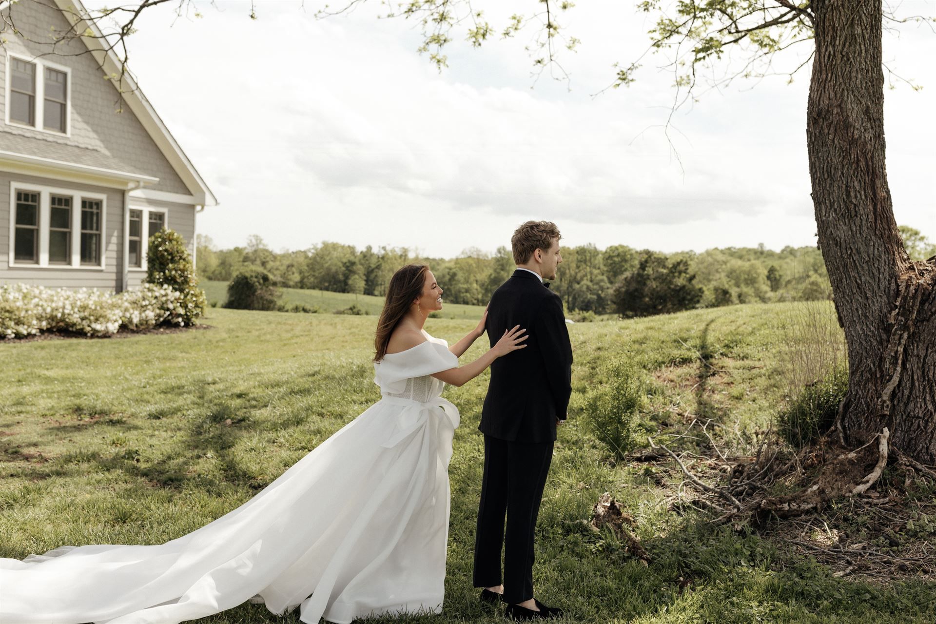 Bride in white gown touches groom's shoulder, standing near tree in front of a house, field in the background.