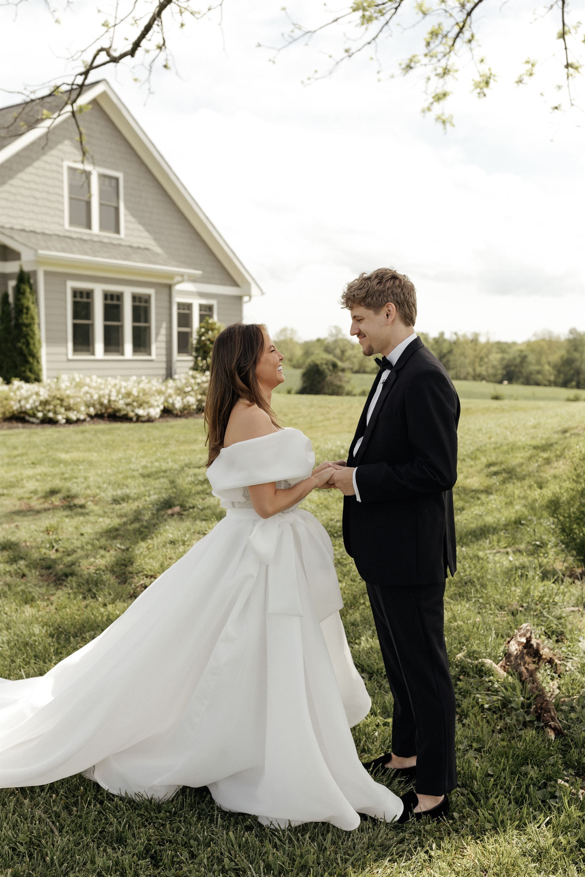 Bride and groom holding hands, facing each other outdoors near a gray house; bride in a white gown.