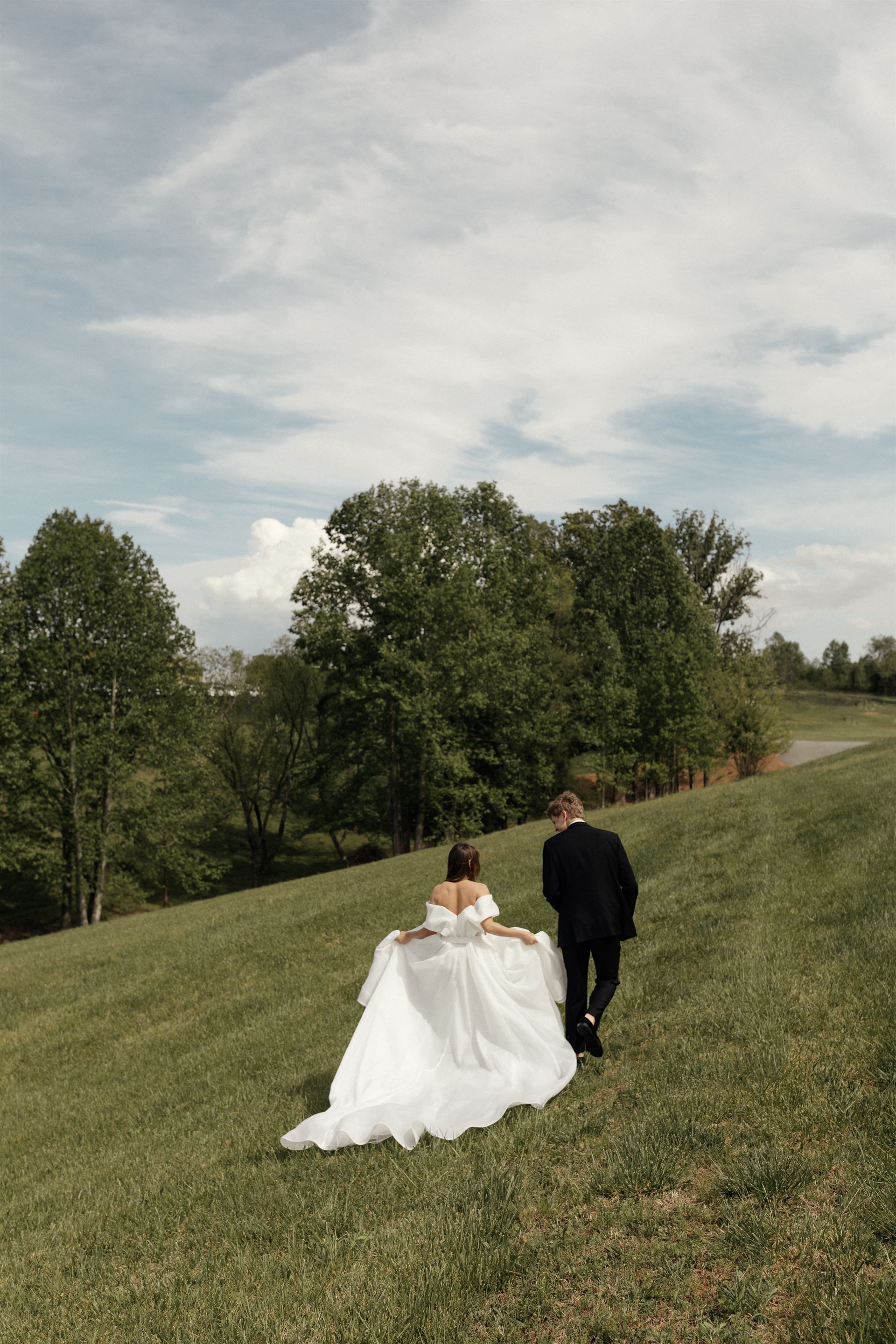 Bride and groom walking uphill on grassy slope; trees and sky in background.