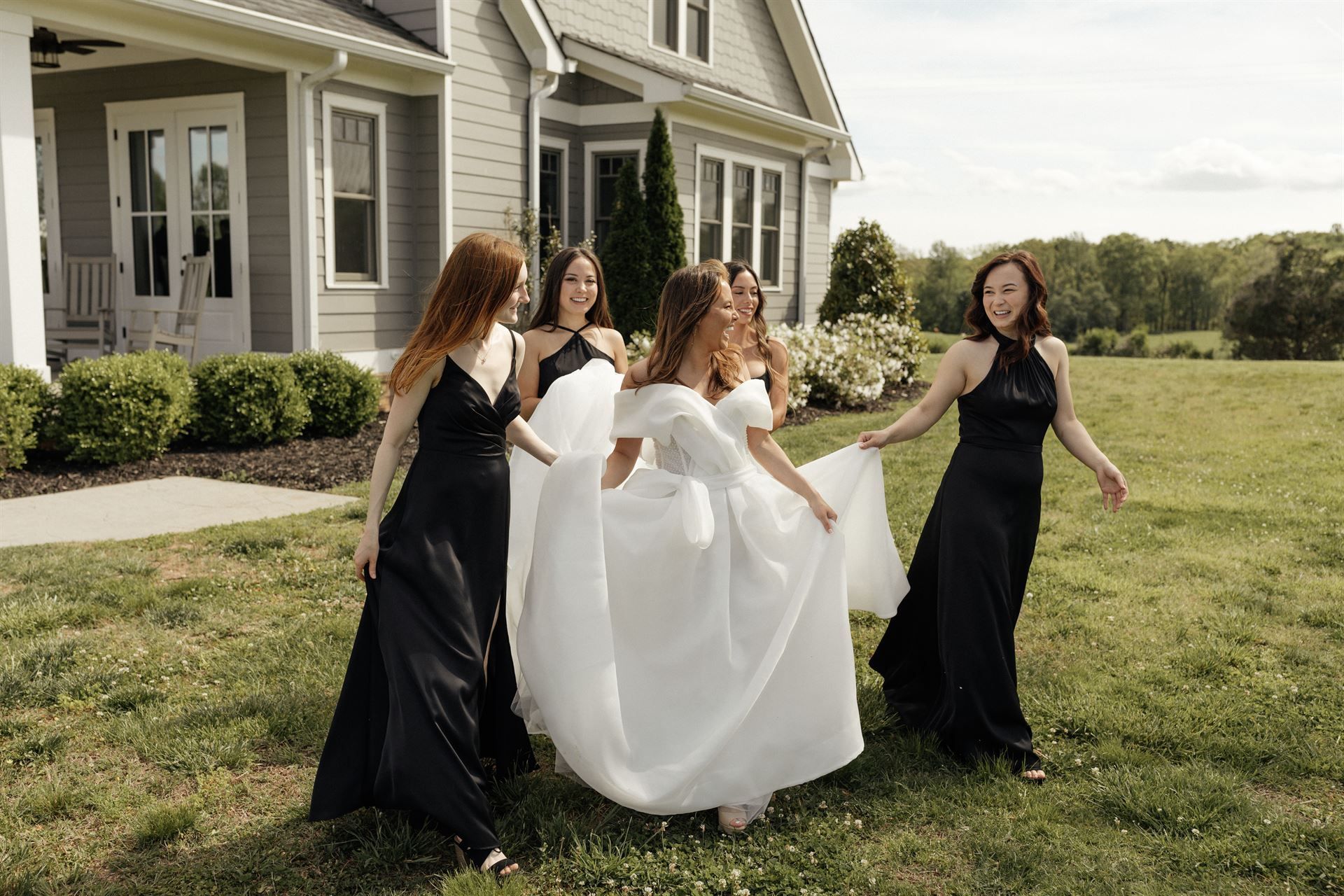 Bride in white dress with bridesmaids in black gowns, in front of a house.