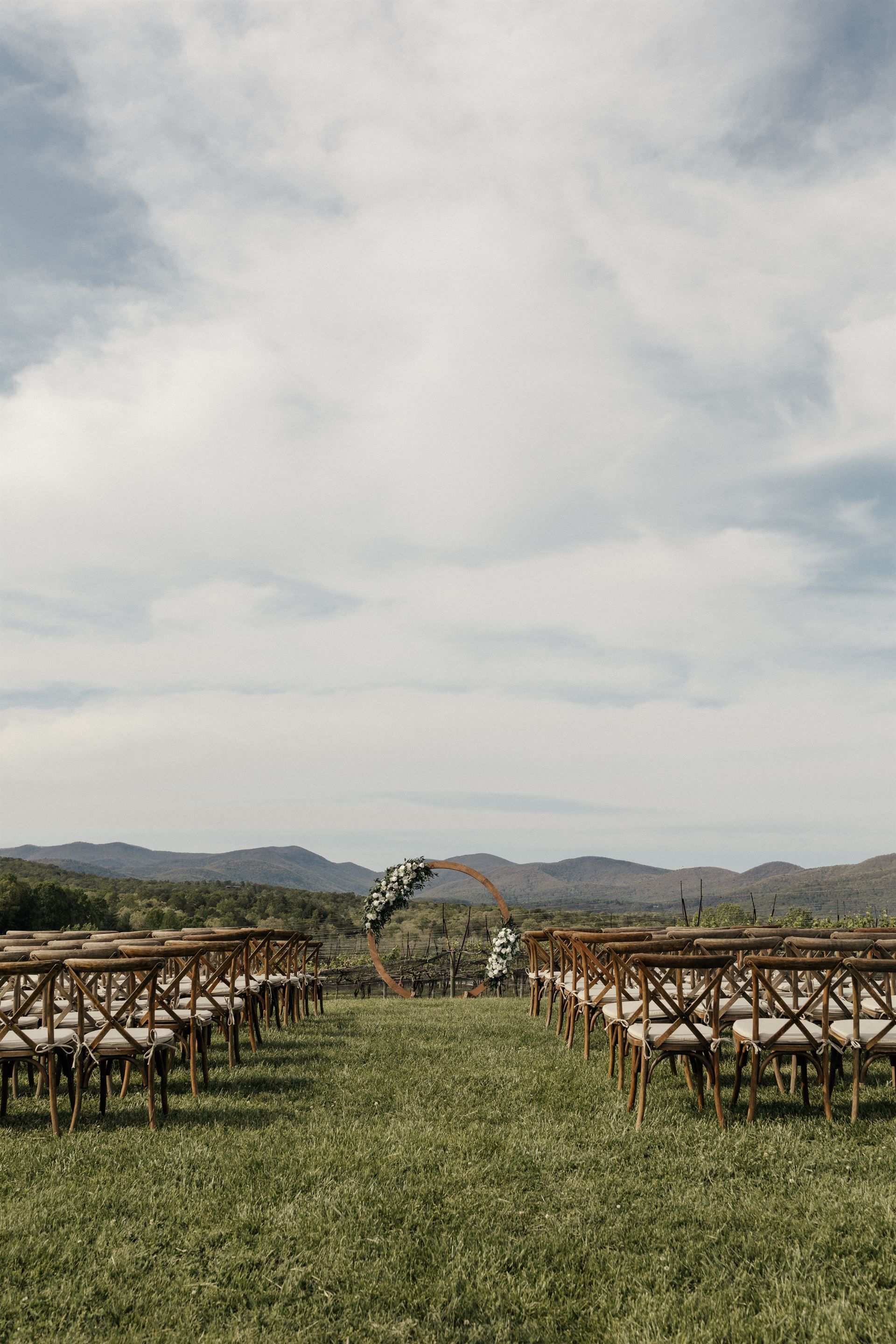 Wedding ceremony setup with chairs facing an arch on a grassy hill overlooking mountains under a cloudy sky.
