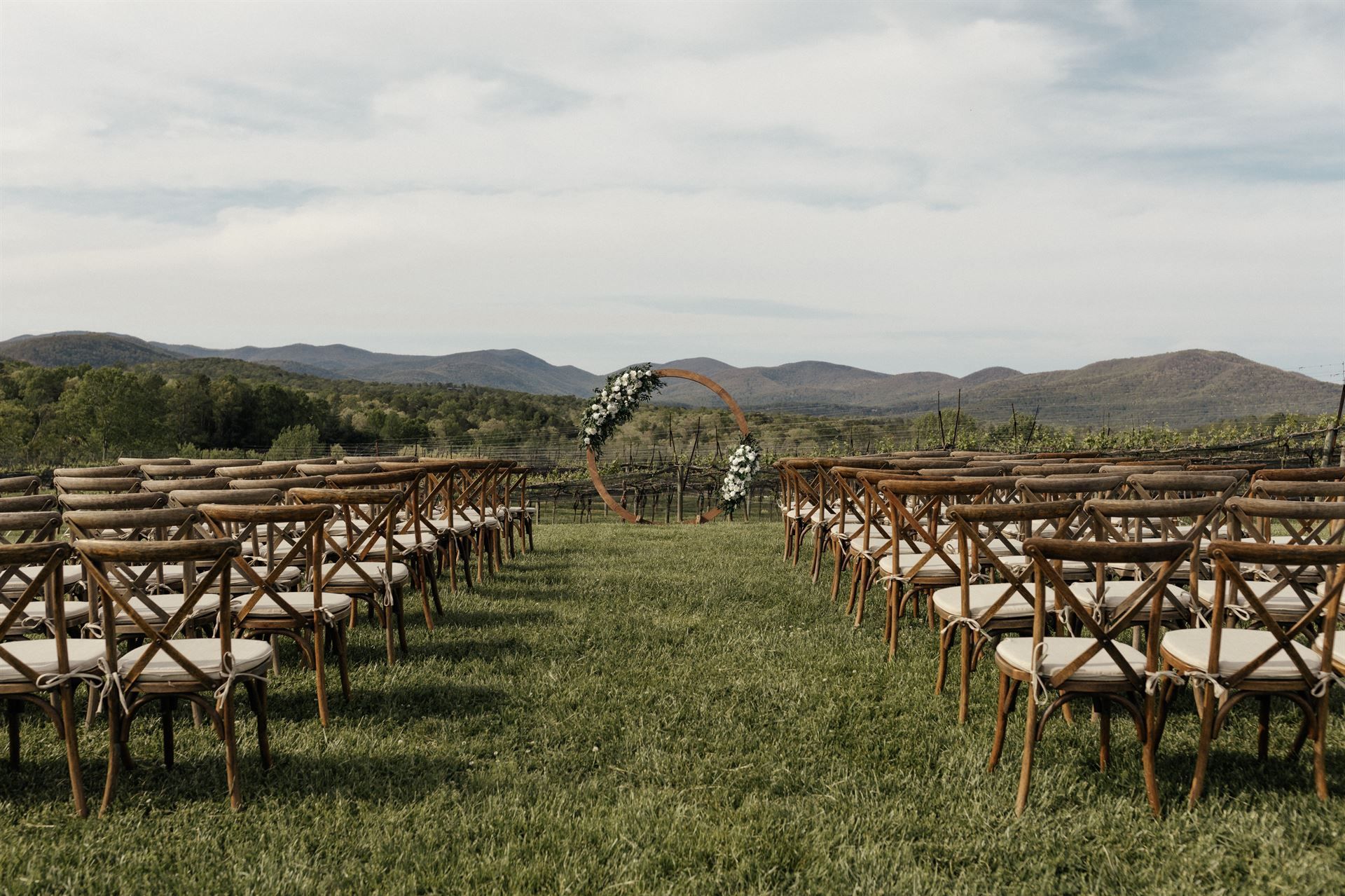 Wedding ceremony setup in field with rows of chairs facing wooden arch, mountains in background.