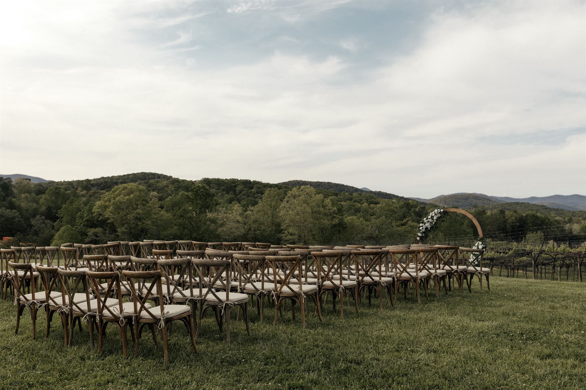 Rows of wooden chairs set up for an outdoor wedding ceremony, with an arch and green landscape in the background.