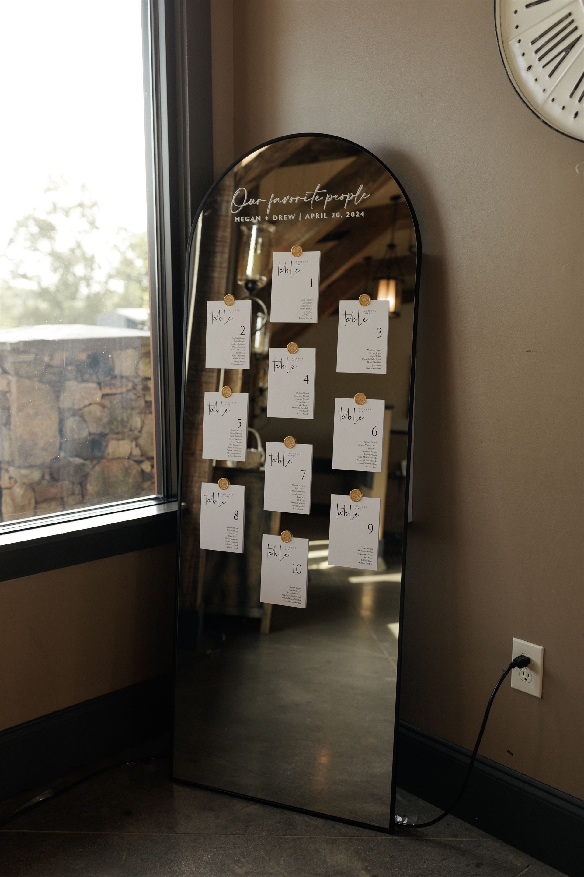Mirror seating chart with guest cards, displayed at a wedding. Brown, neutral tones with window in the background.