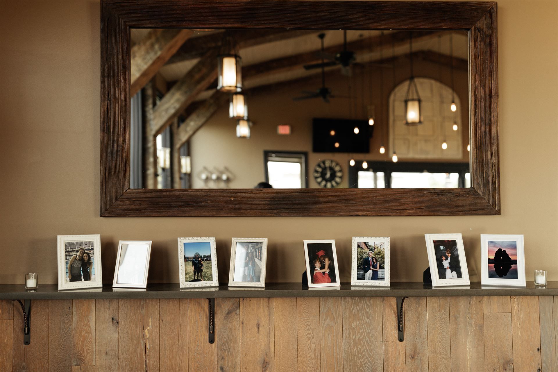Photos in white frames on a wooden shelf beneath a large mirror in a room with exposed beams and hanging lights.