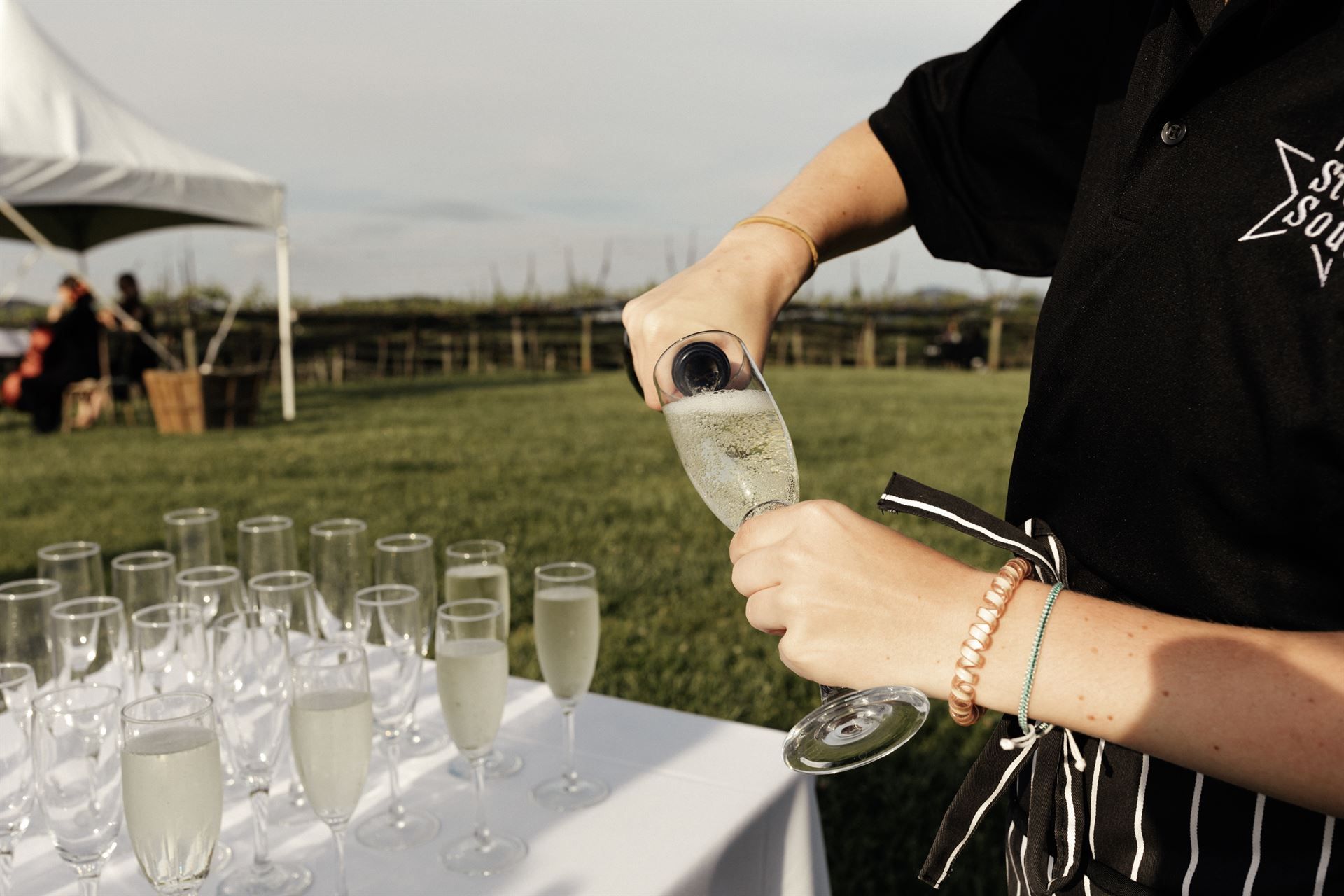 A server pours champagne into a glass at an outdoor event, with filled glasses on a table.